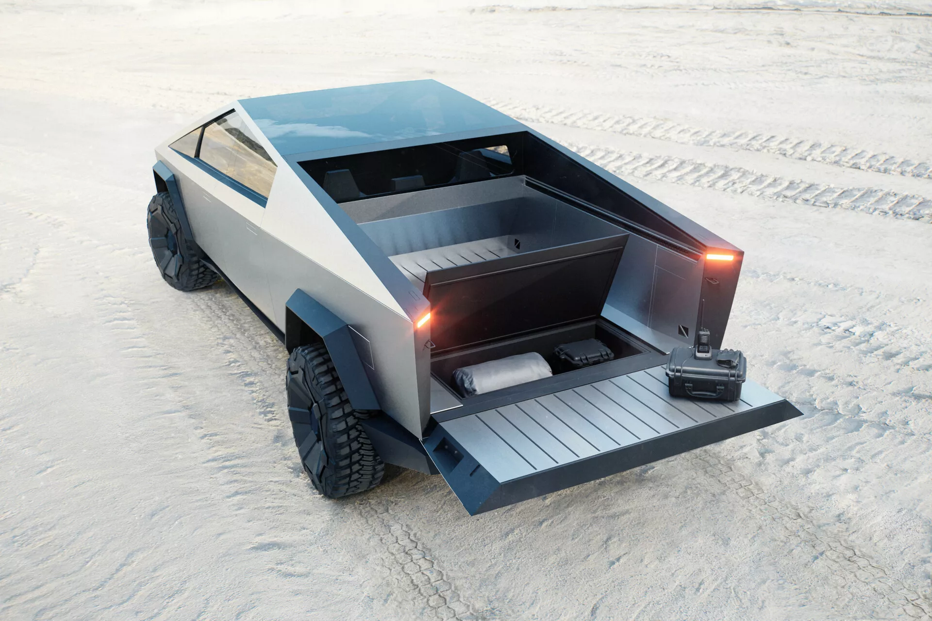 Futuristični Tesla Cybertruck 2021 (1. generacija) parkiran na peščeni površini, posnet z dvignjenega zadnjega tridimenzionalnega pogleda. Kotni, poligonalni dizajn z ravnim tovornjakom in spuščenimi motornimi zadnjimi vrati, ki razkrivajo vgrajeno rampo. Vozilo vključuje tudi zaklenljiv pokrov tovornjaka, velike terenske pnevmatike in mat sivo površino, ki prispeva k njegovi minimalistični, industrijski estetiki. Nobenih tradicionalnih kljuk vrat ali stranskih ogledal ni vidnih, kar nakazuje na umakljive kljuke in ogledala na osnovi kamer. Sumljivo je električno, s poudarkom na praktičnosti in terenskih zmožnostih.