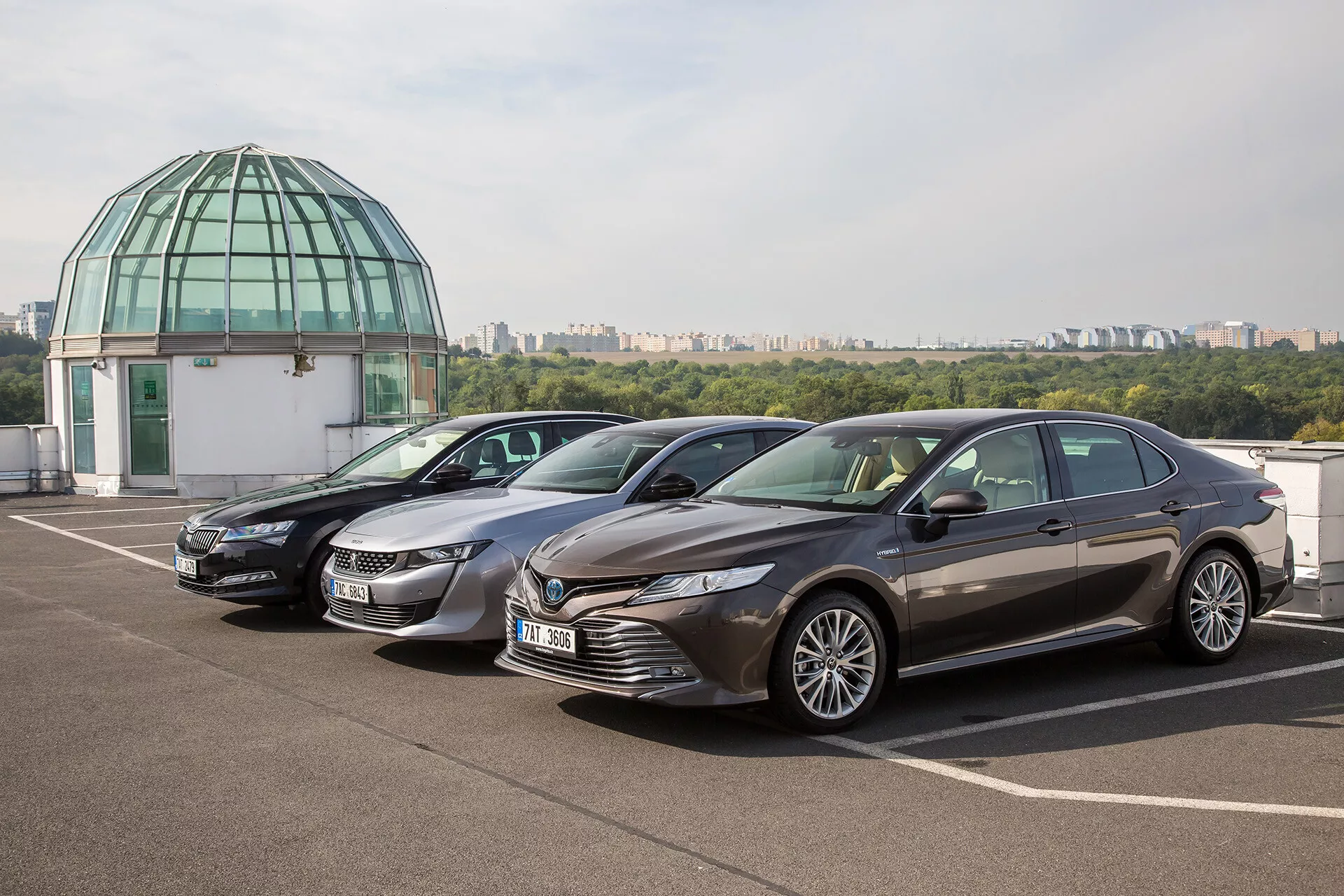 Limuzina Toyota Camry 2019 parkirana na strešnem parkirišču z jasnim nebom in stekleno kupolo v ozadju, predstavlja moderno obliko z dinamično sprednjo rešetko, ostrimi LED žarometi, kiparsko oblikovanim pokrovom motorja in luksuznimi platišči iz zlitine