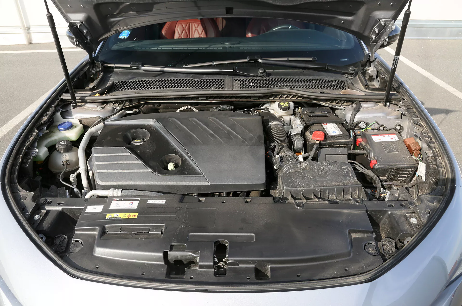 Sprednji pogled na motorni prostor sodobne Toyote Camry 2001 z pokrovom motorja z logotipom, 12-voltno baterijo, zapletenimi žicami in cevmi, posodo hladilne tekočine in pokrovom varovalk, kar kaže na napredno motorno tehnologijo in vzdrževanje.