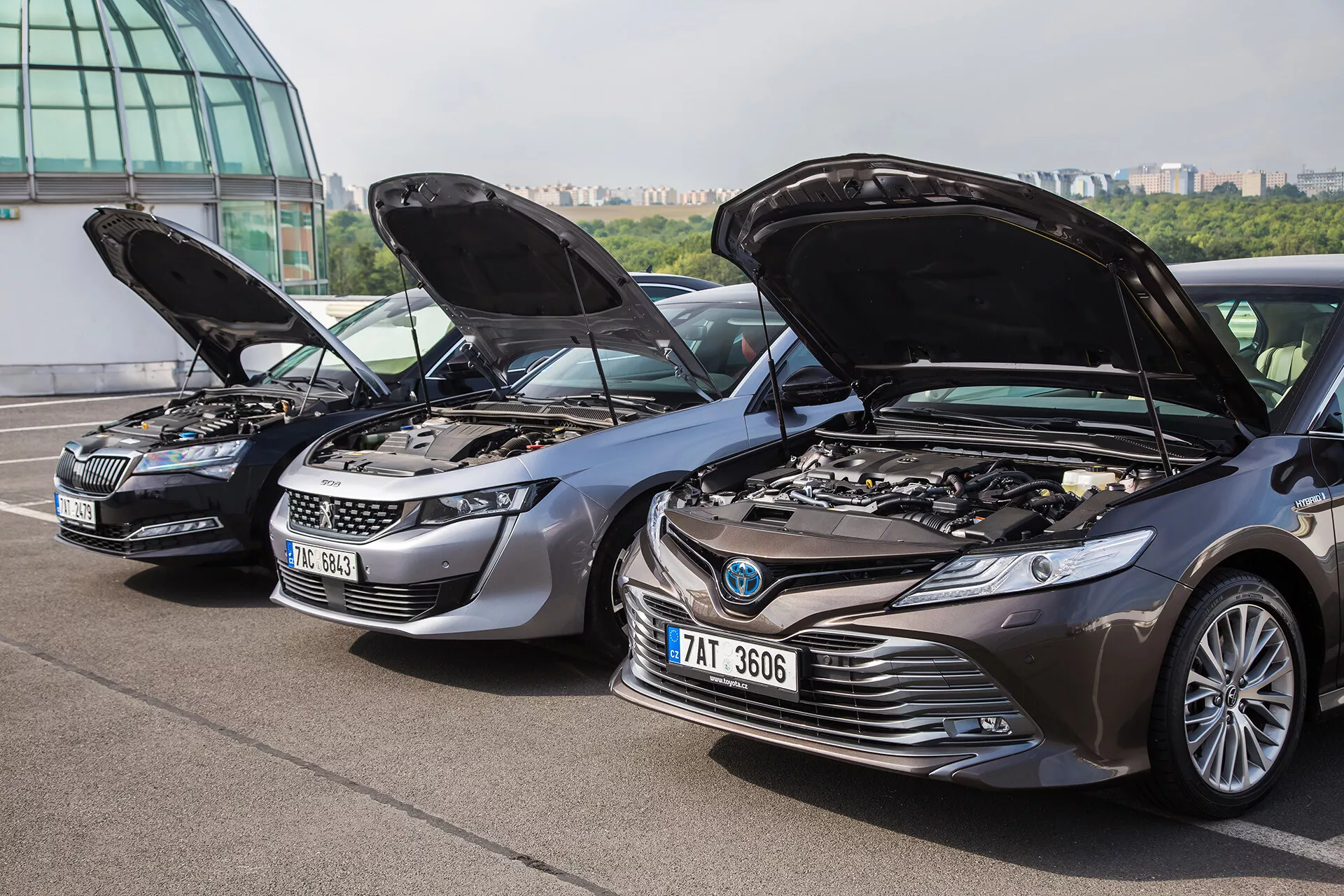 Postavitev treh različnih avtomobilov z odprtimi pokrovi, ki prikazujejo motorje, vključno s srebrno Škodo, sivim SUV-jem Peugeot in temno sivim Toyota Camry, kotni pogled proti mestnemu ozadju s stavbo z zastekljeno fasado in mestnim obzorjem, ki poudarja tehnologijo in oblikovalske značilnosti vozil.