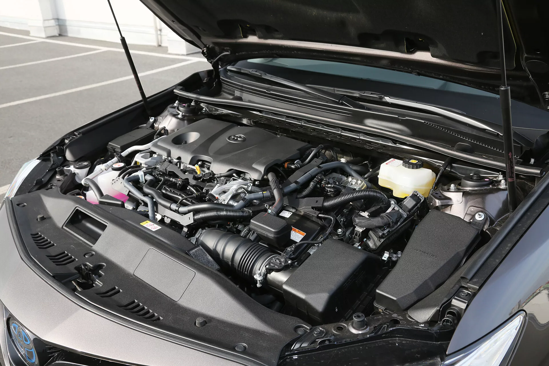 Podroben posnetek motorja sodobnega vozila, ki prikazuje napredne tehnološke komponente, posnet iz rahlo levega srednjega kota s pokrovom motorja odprtim, razkrivajoč podrobnosti motorja in različne rezervoarje sistemov, kar kaže na oblikovanje po letu 2010