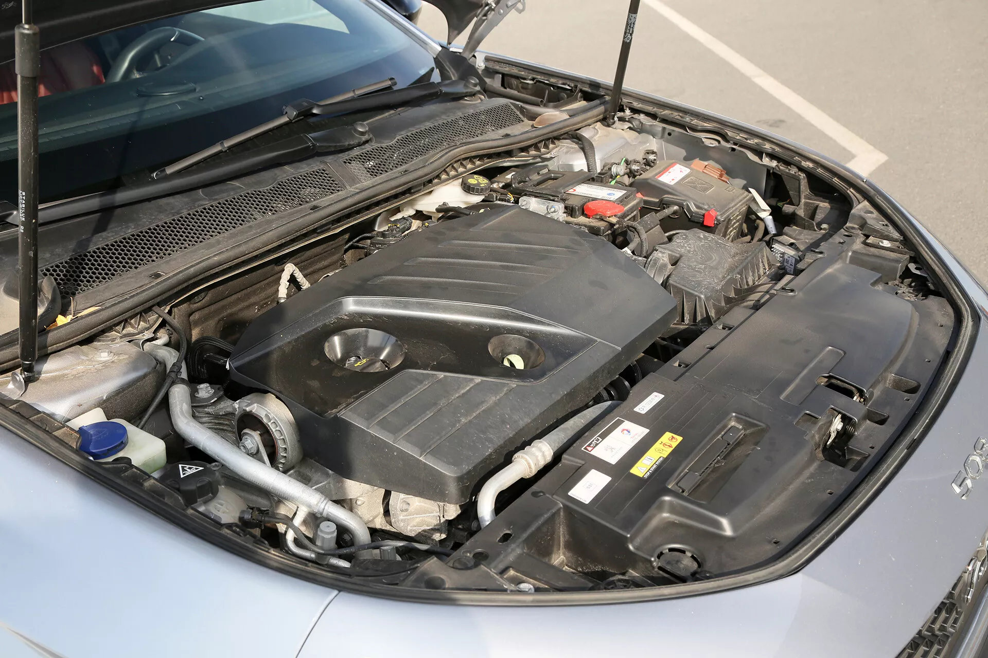 Podroben pogled pod pokrovom limuzine Toyota Camry 2001, ki prikazuje njen motor z jasno vidno baterijo, posodo za hladilno tekočino z modrim zamaškom in različne točke vzdrževanja podnevi z naravno osvetlitvijo