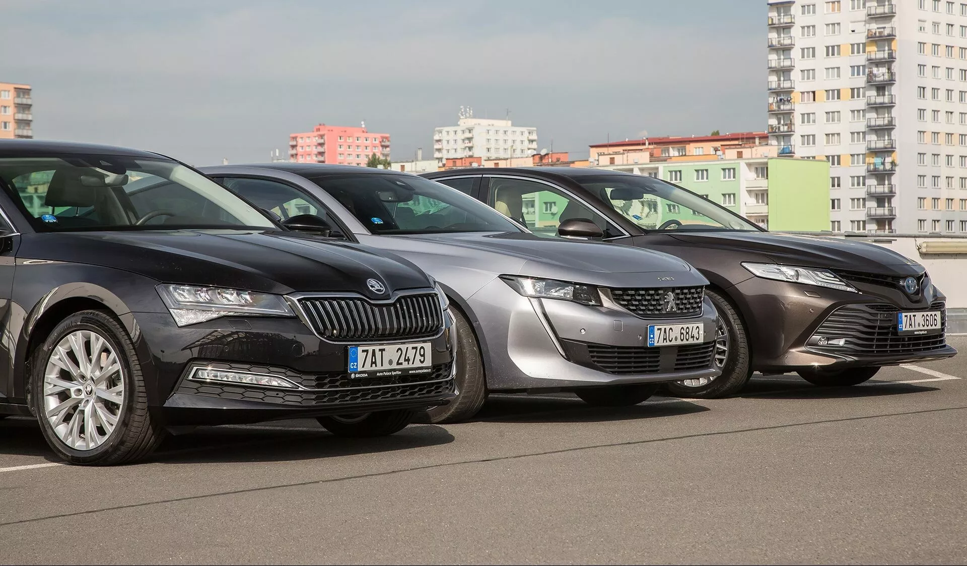 Postavitev Toyote Camry 2019, s temnimi, srebrnimi in sivimi modeli v mestnem okolju, ki prikazuje pogled od spredaj in del strani z LED žarometi in elegantnimi oblikovnimi elementi