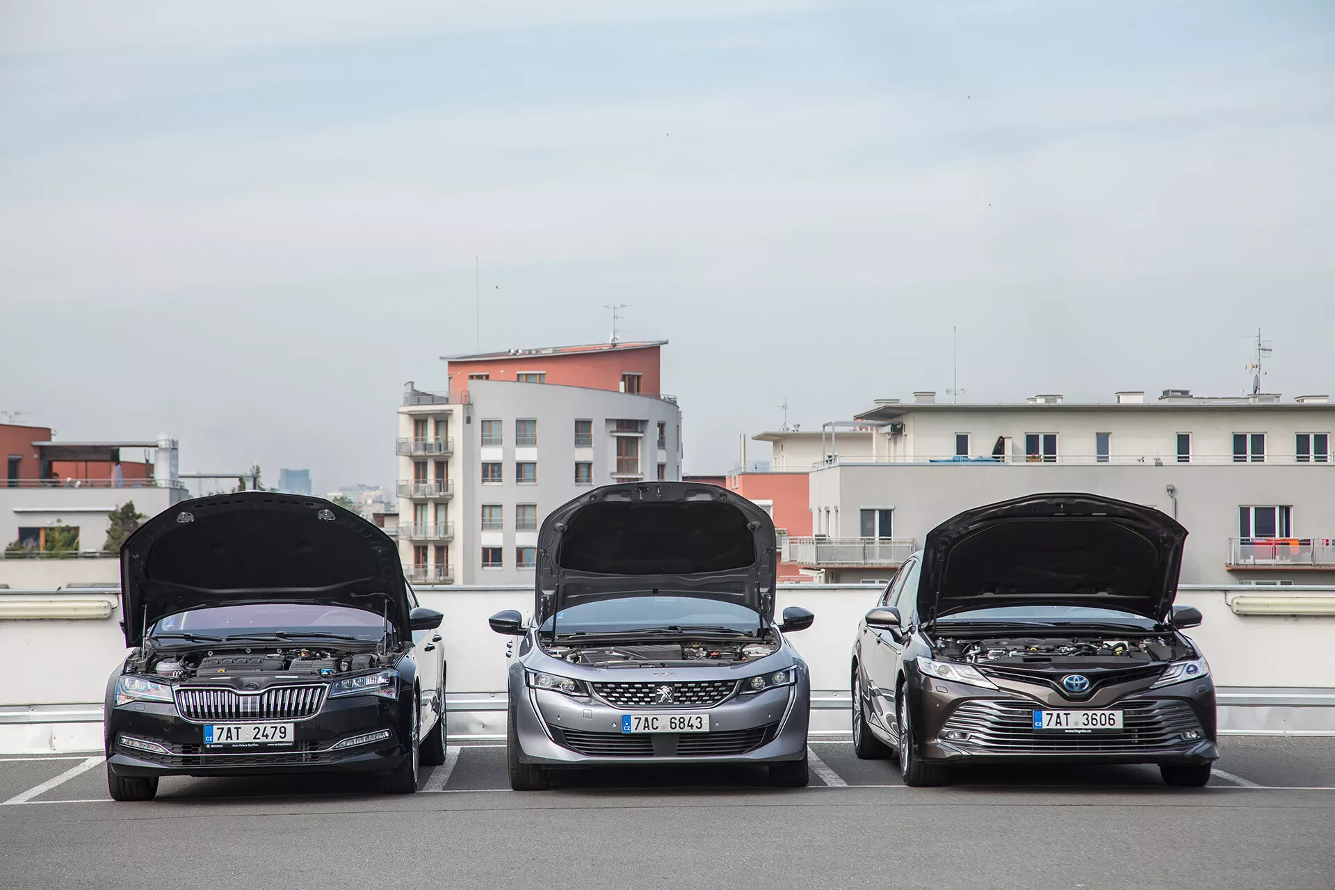 Tri limuzine Toyota Camry letnik 2001 parkirane ena ob drugi na strešnem parkirišču, čelni pogled z odprtimi pokrovi razkriva motorne prostore, dnevna svetloba prikazuje tehnološke funkcije in oblikovanje na ozadju mesta