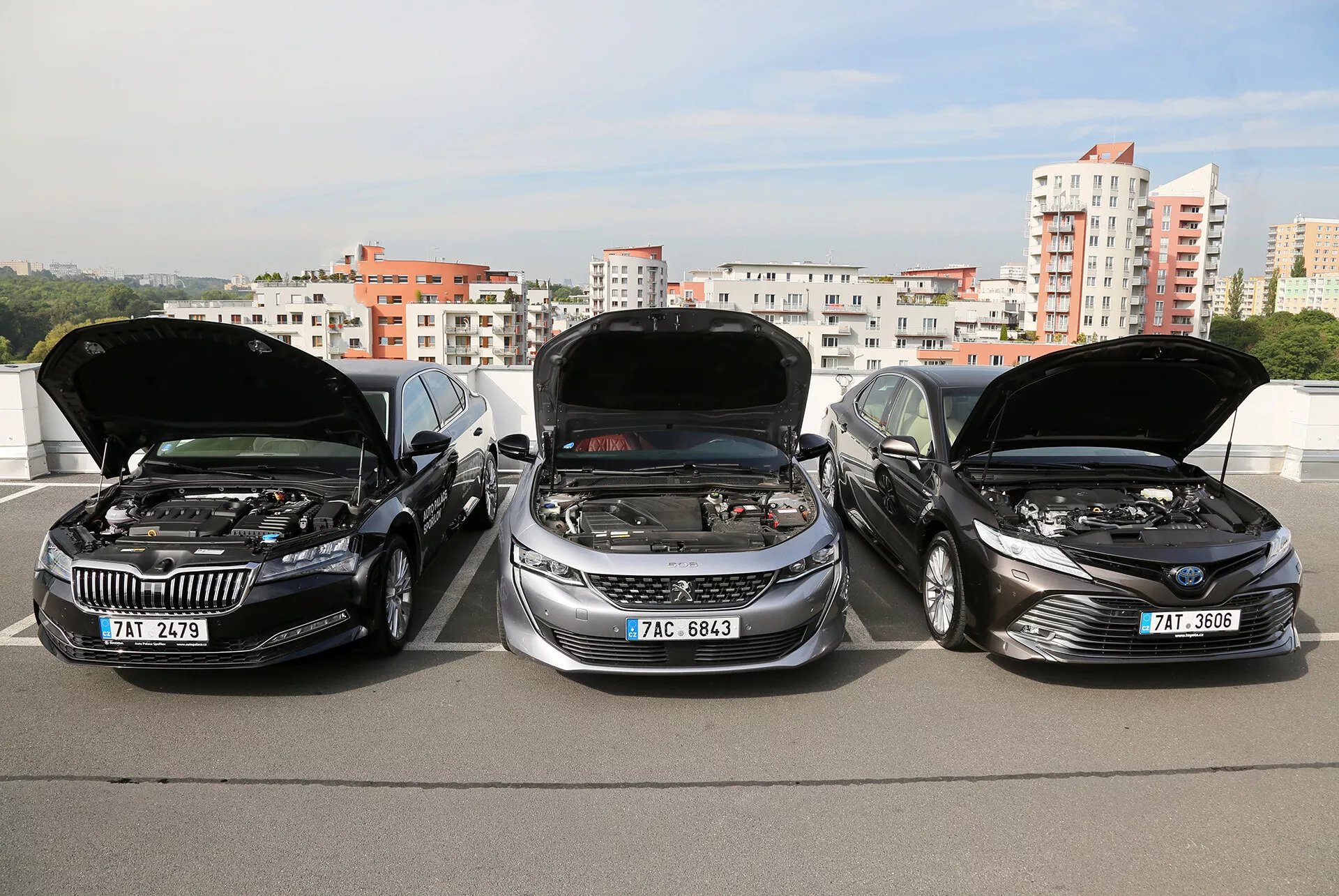 Modeli limuzine Toyota Camry 2019 z odprtimi pokrovi, ki prikazujejo motorje, parkirani drug ob drugem na dvignjeni konstrukciji, z mestno obzorje v ozadju, kar poudarja elegantne, sodobne oblikovanje vozil in napredno tehnologijo motorja.