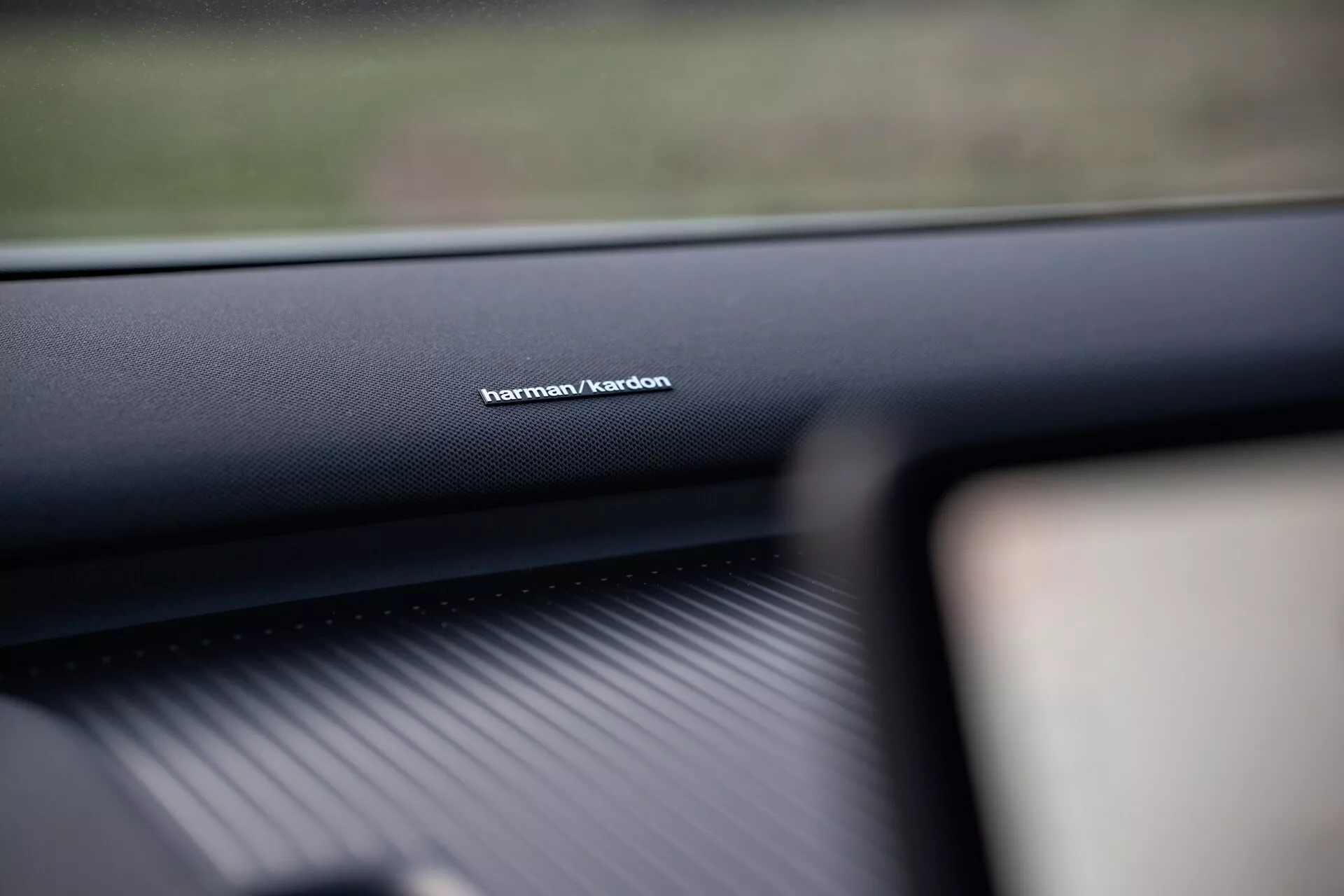 Notranji posnetek Volva EX30 2024, osredotočen na blagovno znamko avdio sistema Harman/Kardon na armaturni plošči na strani sovoznika, ki prikazuje kakovosten material in poudarja vrhunsko tehnologijo zvočnega sistema.