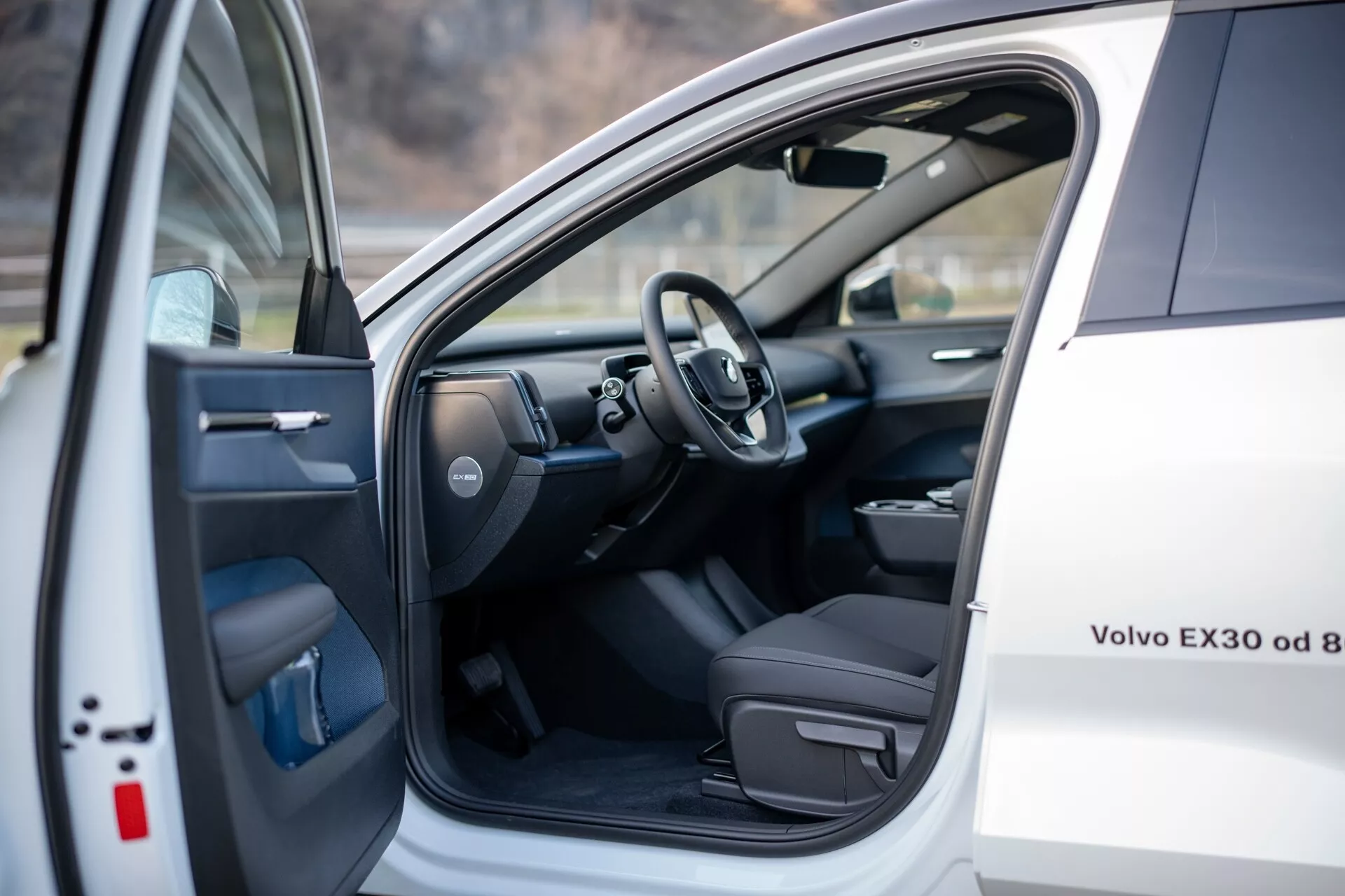 Pogled na notranjost bele Volva EX30 2024, ki prikazuje voznikov sedež in področje kokpita z odprtimi sprednjimi levimi vrati z električnimi kontrolami in pogled na volan ter del armaturne plošče