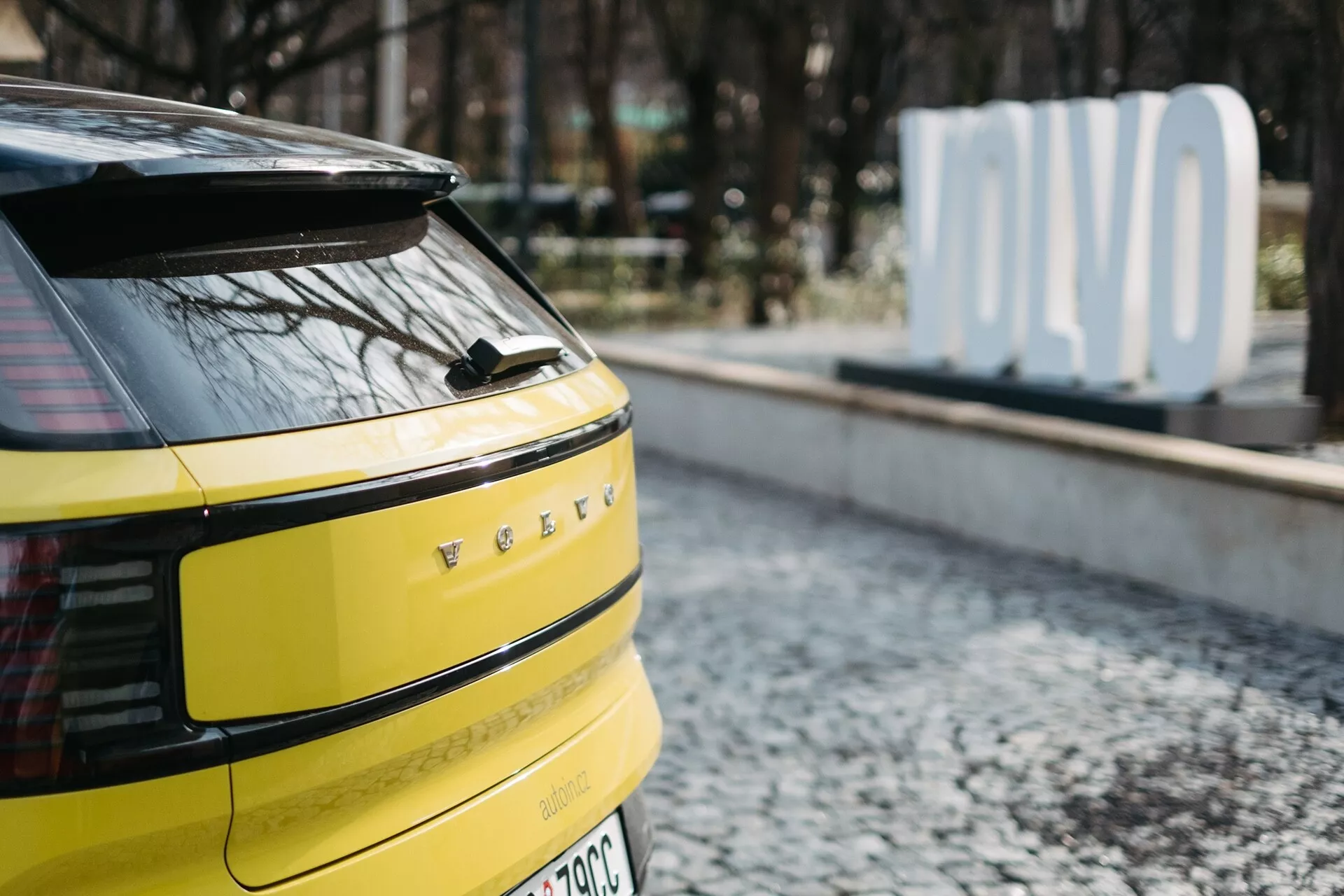 Prvi načrt zadnjega dela svetlo rumenega Volvo EX30 2024 z vodoravnimi LED zadnjimi lučmi, morebitno zadnjo kamero ali senzorji, enim zadnjim brisalcem, oznako elektro vozila ali hibrida in anteno v obliki morskega psa, parkiran na tlakovani površini z zamegljenim znakom 'OLVO' v ozadju