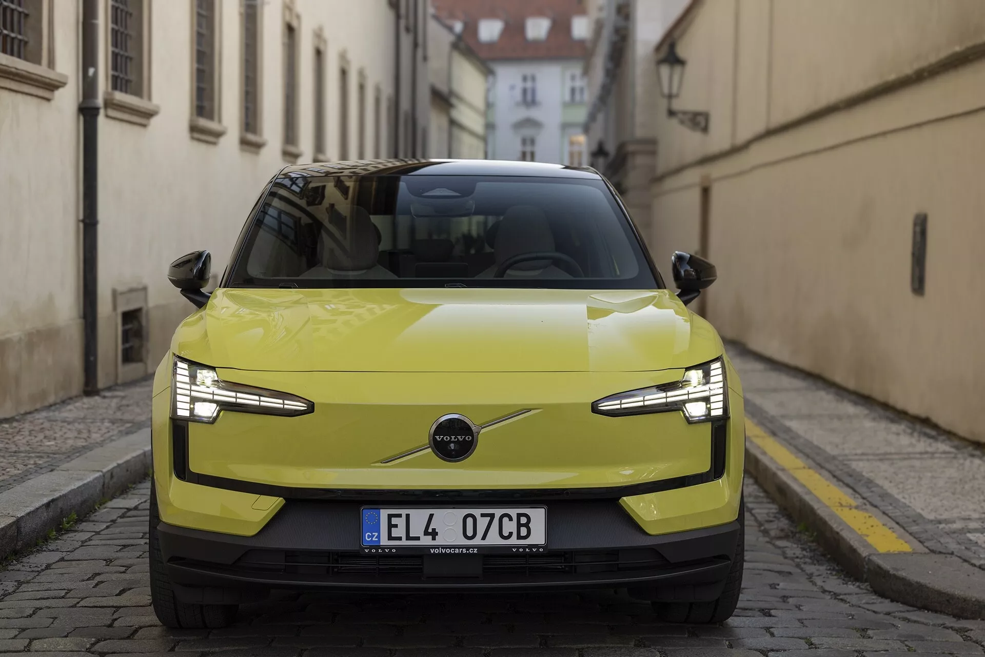 Sprednji pogled na Volvo EX30 2024 v limetno rumeni barvi, ki poudarja njegove elegantne LED žaromete, minimalistično rešetko, ki nakazuje na električni pogon, in napredne tehnološke značilnosti, kot so senzorji parkiranja in morebitni ADAS, na ozadju zgodovinske evropske ulice