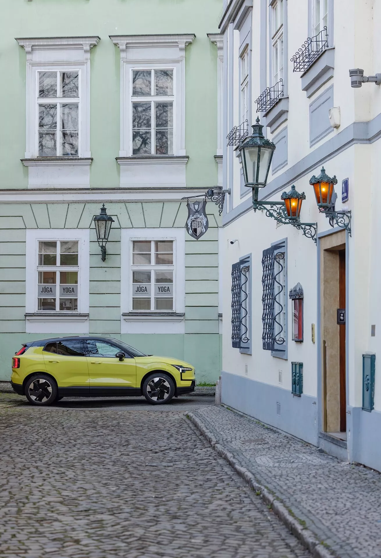 Volvo EX30 2024 parkiran na tlakovani ulici v starem mestnem jedru, posnet s tročetrtinskega zadnjega kota, ki prikazuje desno stran in zadnji del avtomobila, vključno s kolesi, zadnjimi lučmi in delnimi podrobnostmi strehe v svetlo rumeni barvi