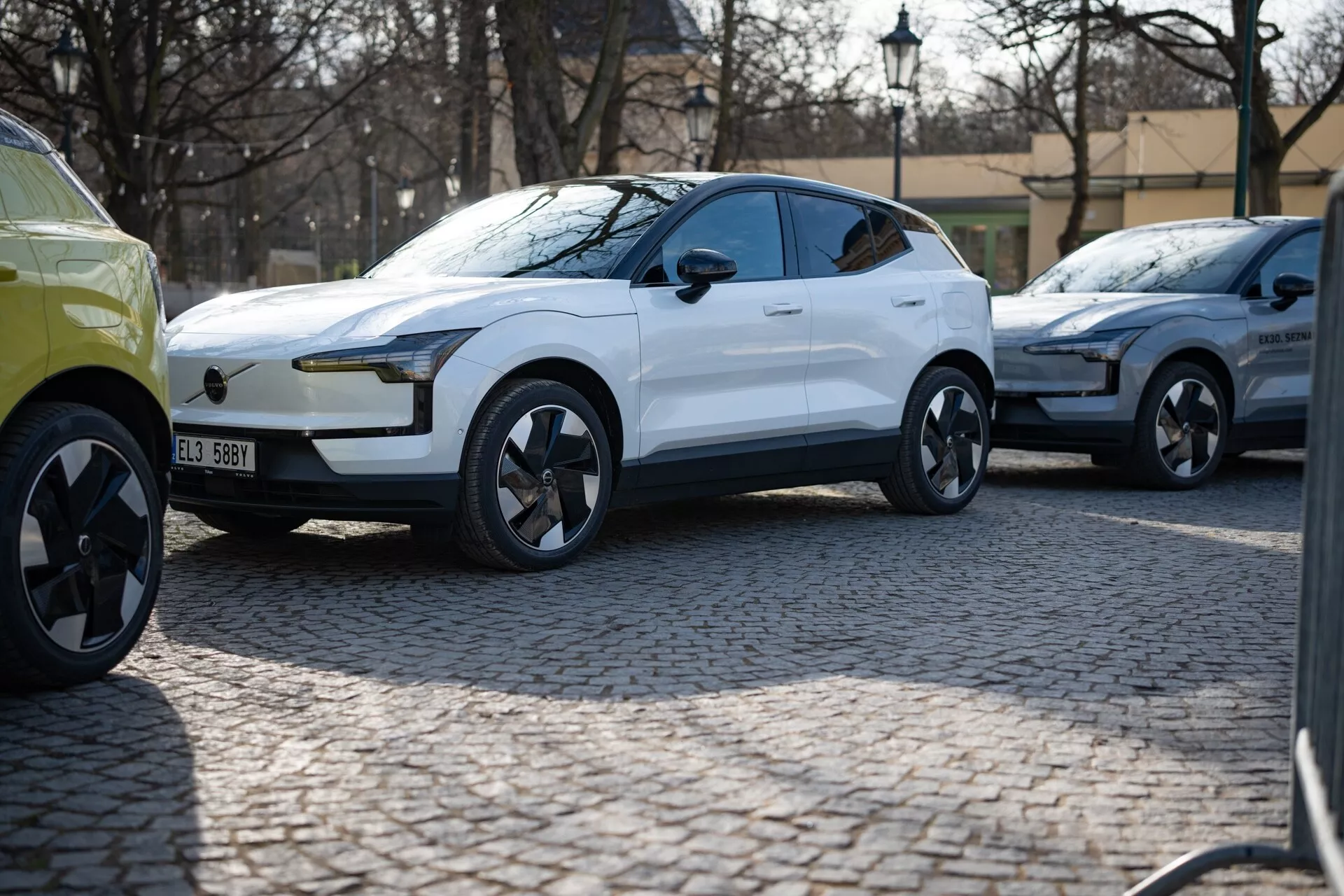 Beli moderni crossover SUV 2024 v tričetrtinskem pogledu parkiran na tlakovani ulici, z sijočo barvo, LED dnevnimi lučmi, vratnimi kljukami v liniji z obliko in dinamično oblikovanimi liti kolesi, ki sugerirajo luksuz in zmogljivost, verjetno električno vozilo.