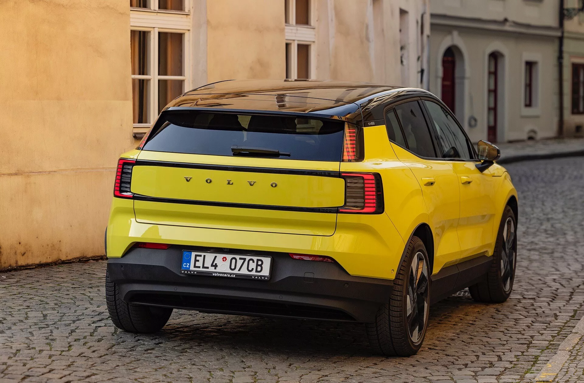 Rumeni SUV Volvo EX30 2024 prikazuje svojo značilno ukrivljenost zadnjega blatnika, LED zadnje luči in varnostne funkcije, parkiran na tlakovani ulici s pogleda s treh četrtin zadaj