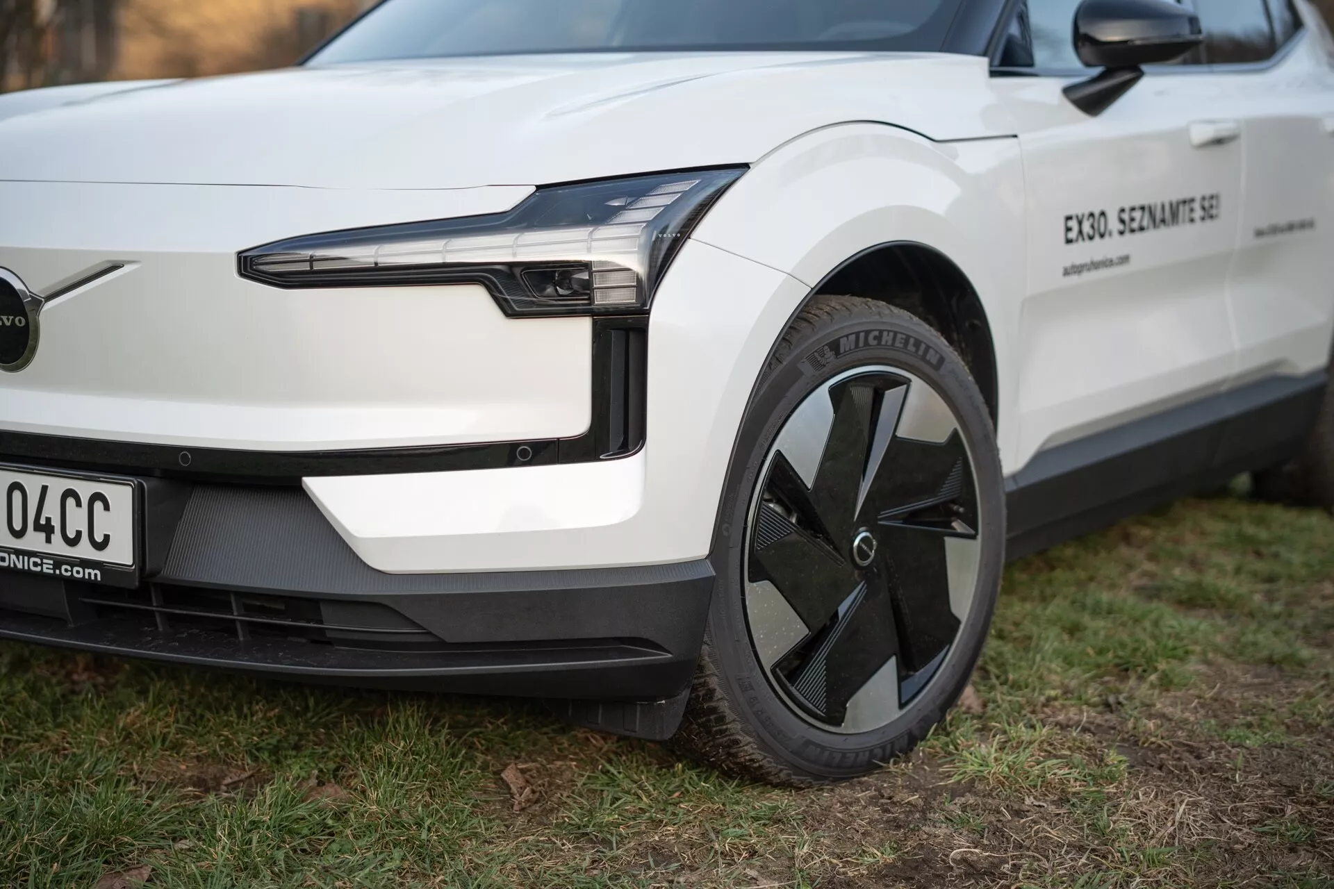 Podroben pogled na električno vozilo Volvo EX30 2024 iz perspektive treh četrtin, ki poudarja njegovo LED žaromet, aerodinamično zasnovo kolesa in minimalistični sprednji odbijač brez rešetke, v beli barvi s črnimi obrobami.