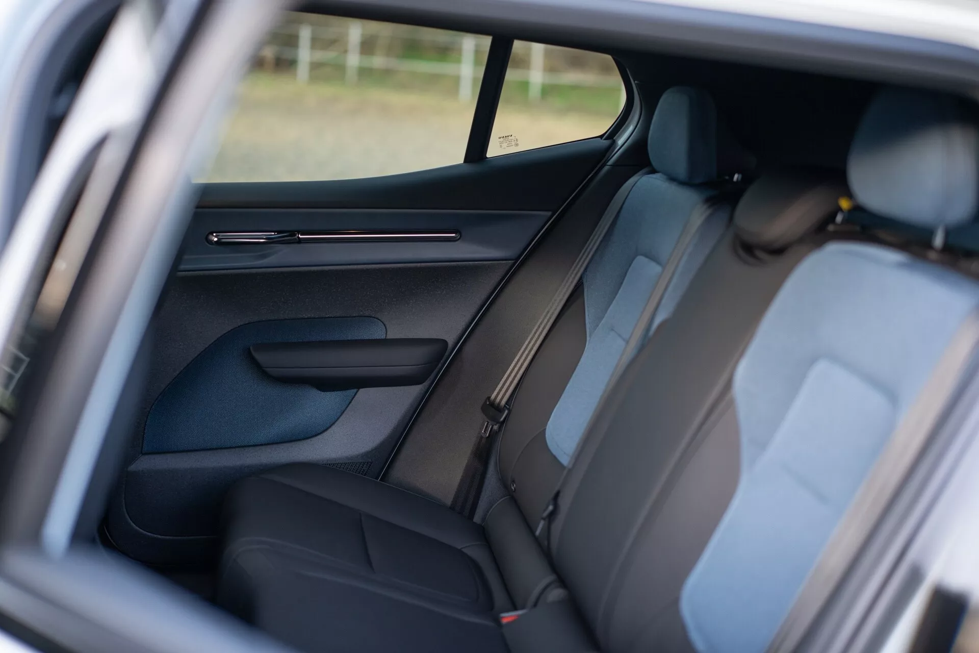 Notranjost Volvo EX30 2024 prikazuje zadnji potniški del skozi odprta vrata sprednjega sopotnika, značilnosti temno oblogo, vidne naslonjale za glavo, podrobno oblikovan panel vrat z upravljalnimi elementi, B-stebriček design, varnostne pasove za zadnje sedeže, delno vidno stransko okno, ki izboljšuje vzdušje v notranjosti, in poudarek na razporeditvi sedežev za udobje in varnost potnikov.