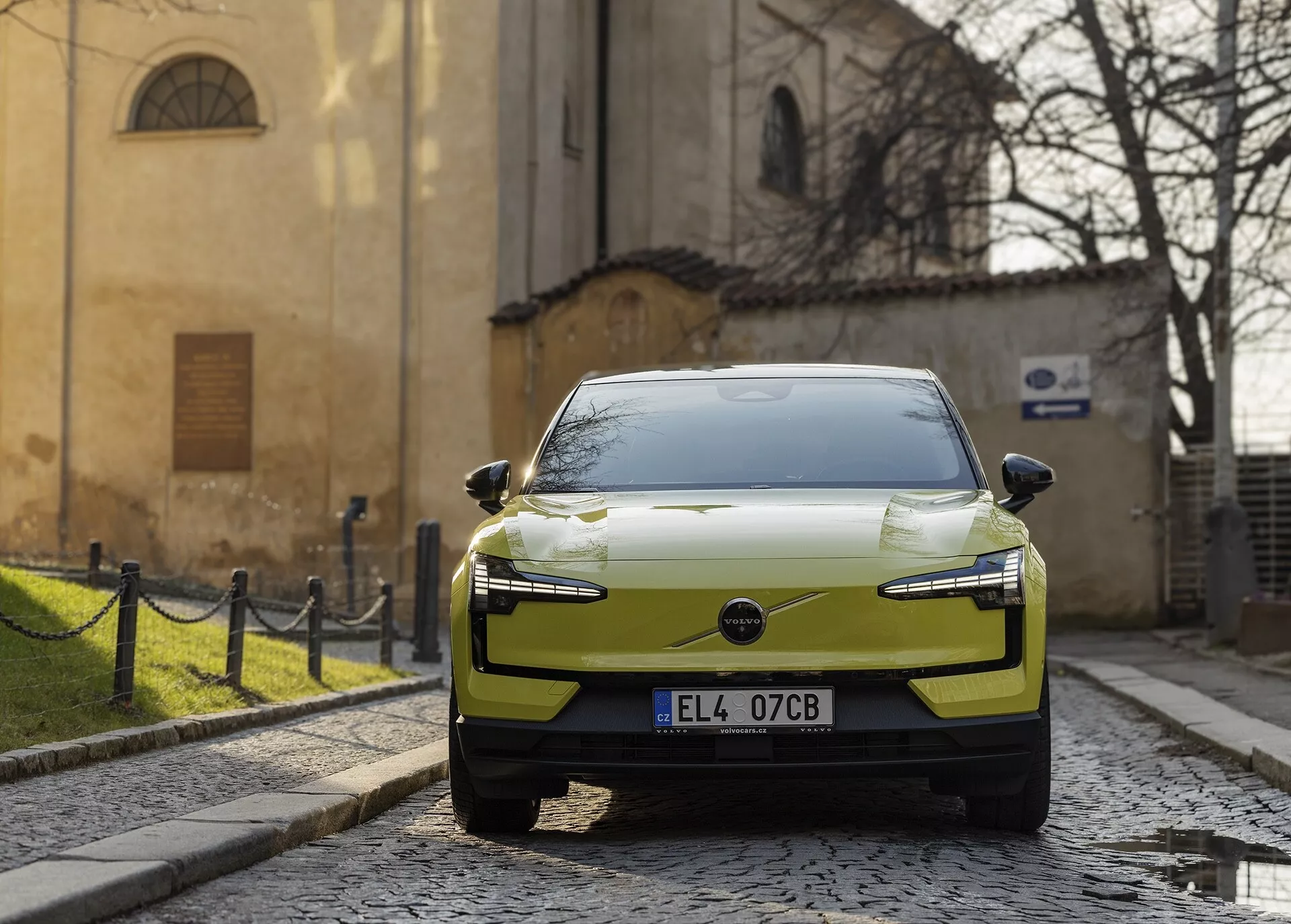 Moderen električni avtomobil Volvo EX30 letnik 2024 v odtenku chartreuse parkiran na tlakovani ulici, pogled iz desno-sprednje perspektive z LED žarometi in aerodinamičnim dizajnom pokrova motorja izstopa na zgodovinskem evropskem ozadju.