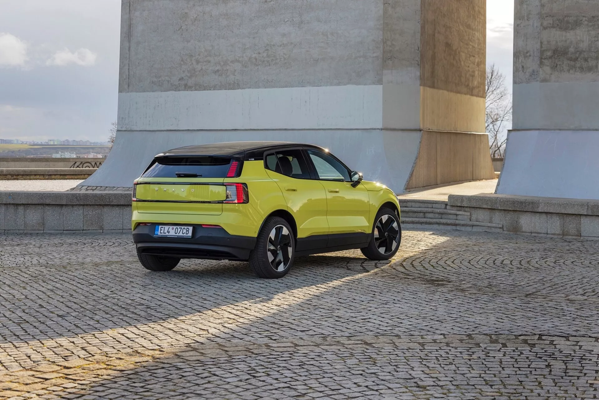 Limetno zeleni SUV Volvo EX30 2024 v pogledu z zadaj treh četrtin, ki prikazuje stranski profil, zadnje luči, prtljažna vrata in črna lita platišča ob kamnitih zidovih na tlakovanem tleh