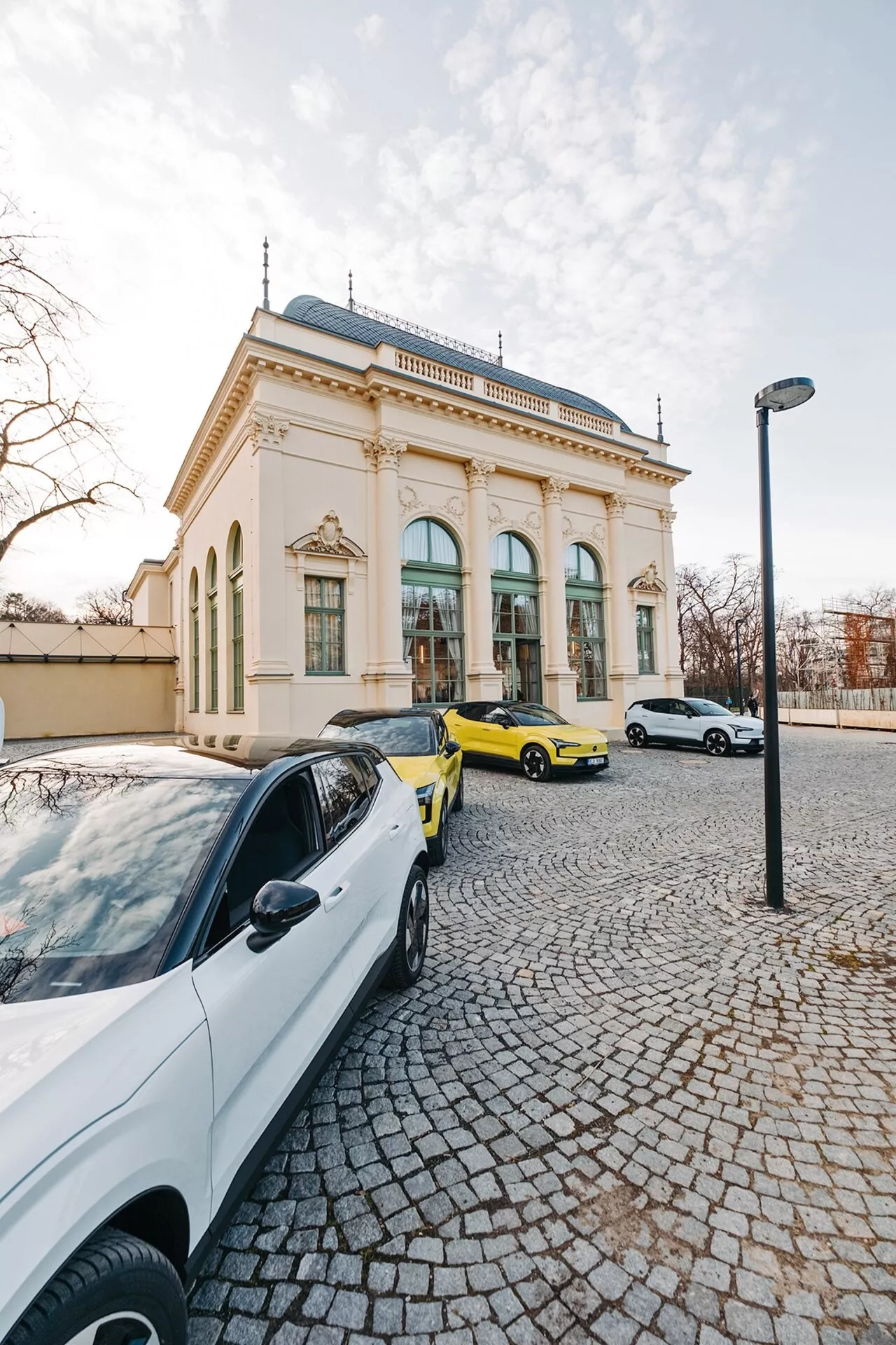 Ulični prizor s tremi avtomobili in elegantno zgradbo v ozadju, posnet z širokokotne perspektive, ki poudarja beli sodobni sedan, živahno rumeni športni avto in beli sedan ali hatchback v svetlobi poznega popoldneva