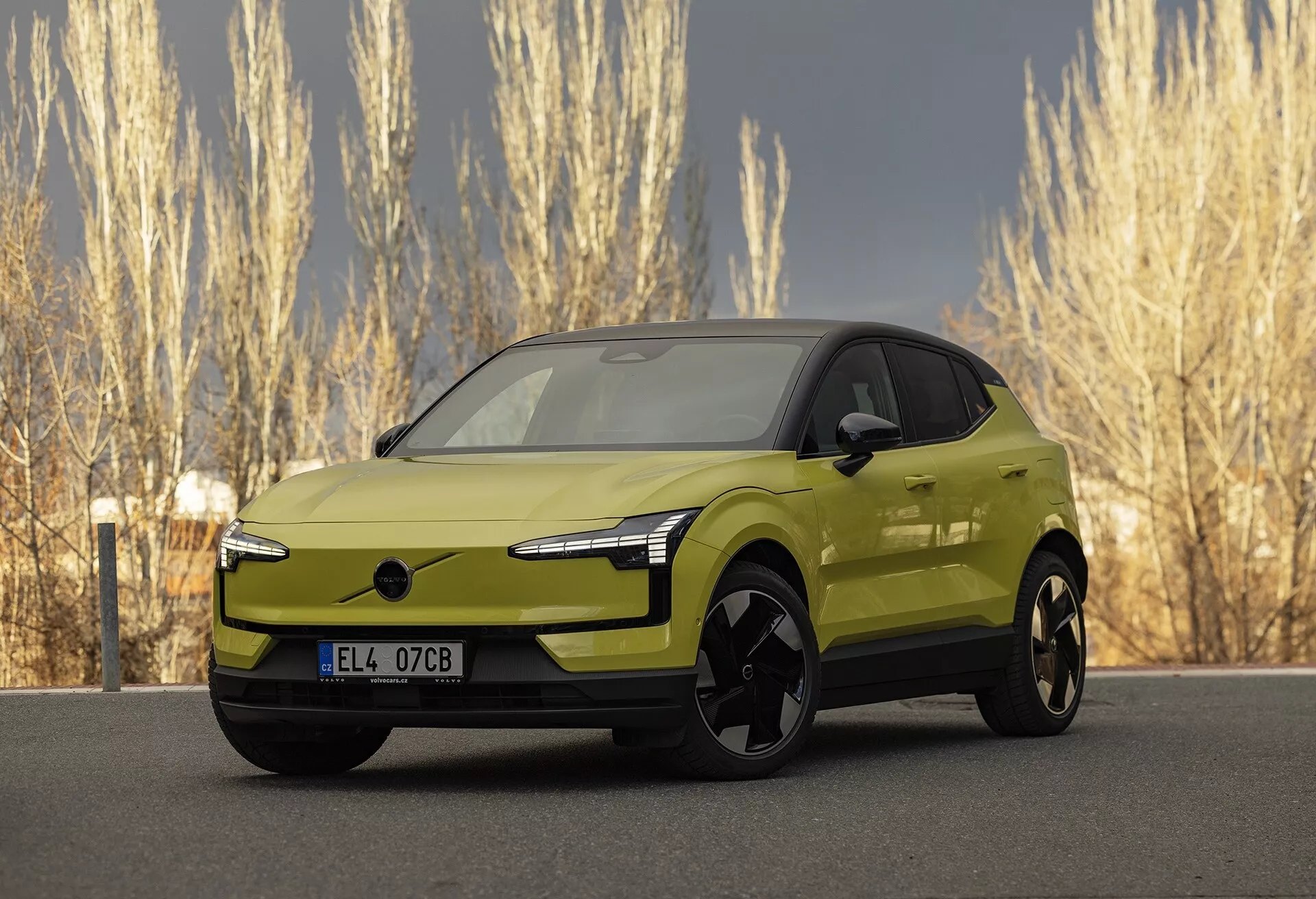 Moderen električni avtomobil Volvo EX30 2024 v apneni zeleni barvi v kotnem sprednjem pogledu, ki poudarja njegove elegantne LED luči, pomanjkanje tradicionalne rešetke za izboljšanje aerodinamike, velika črna lita platišča, zunanja ogledala v barvi karoserije z morebitno integrirano tehnologijo, gladki in aerodinamični stranski profil s podobo kupelista in živo metalno končno obdelavo, parkirano v naravnem okolju