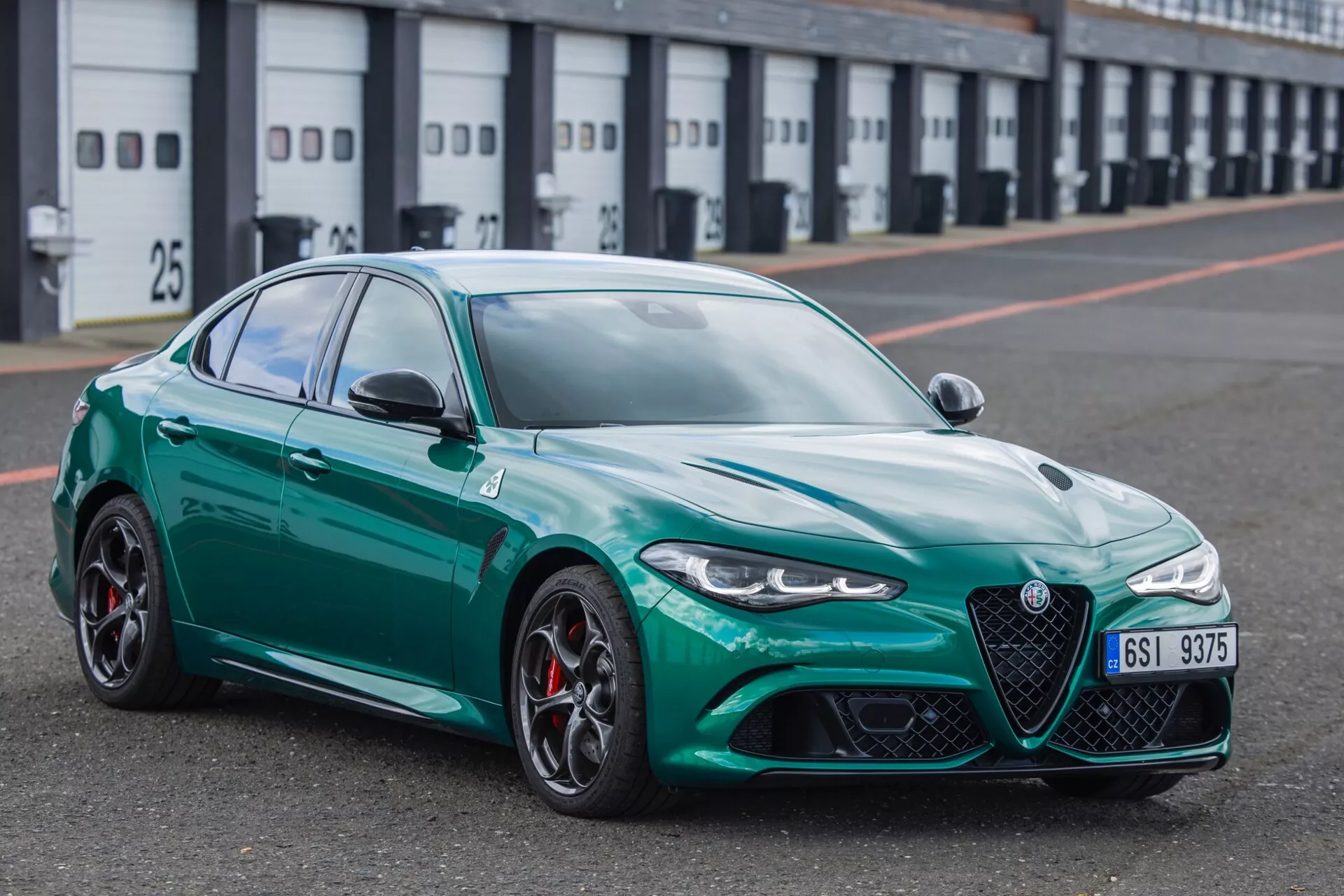 Športna limuzina Alfa Romeo Giulia Quadrifoglio 2023 v metalni finiš, videna iz prednjega kota tri četrt, na zaustavnem pasu dirkališča, prikazuje svojo značilno sprednjo rešetko, elegantne LED žaromete, velike zračne vhode, lita kolesa z večkrakim dizajnom z rdečimi zavornimi čeljustmi, športni aerodinamični profil, napredno obliko stranskih ogledal in vzorec tekalne plasti pnevmatik usmerjen v zmogljivost.