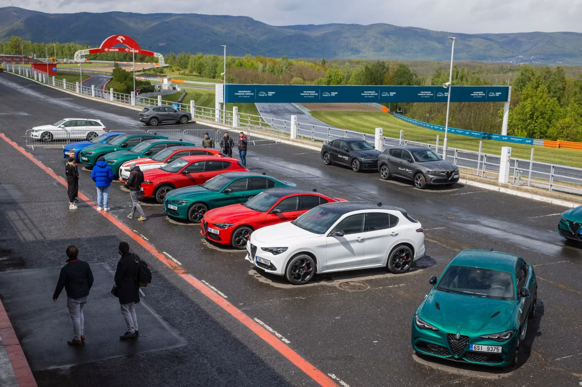 Alfa Romeo Giulia Quadrifoglio leta 2023 na slikovitem dirkališču, prikazuje elegantno aerodinamično obliko med vrsto športnih in luksuznih vozil pod delno oblačnim nebom