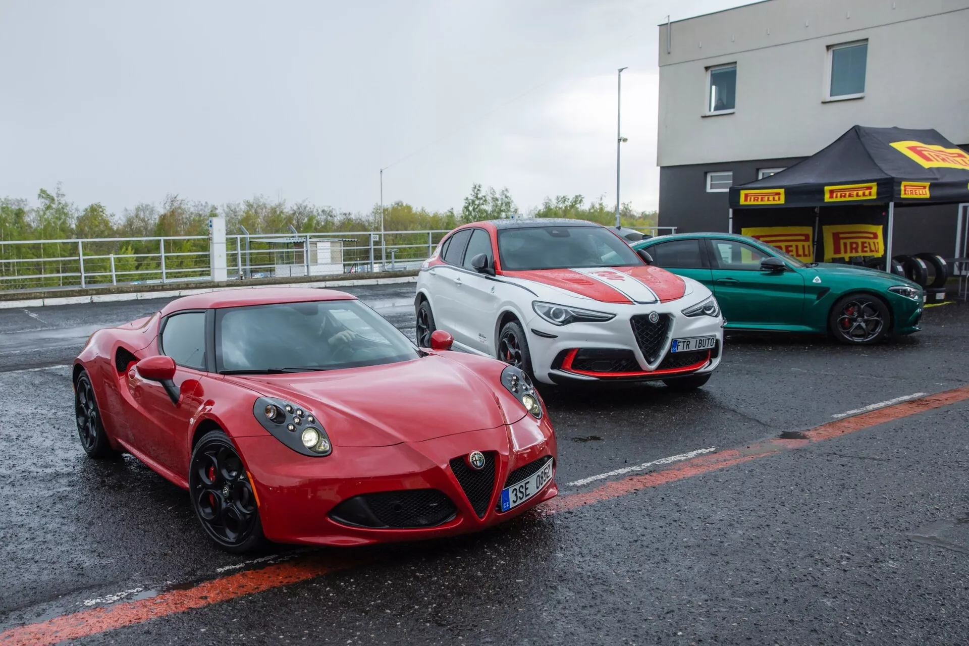 Alfa-Romeo Giulia Quadrifoglio 2023, rdeče športno kupe parkirano v boksih dirkališča pod oblačnim nebom, s prikazom sprednjega in stranskega profila, LED žarometi, trikotna sprednja rešetka z logotipom, veliki zračniki na odbijaču in zlitinska kolesa.