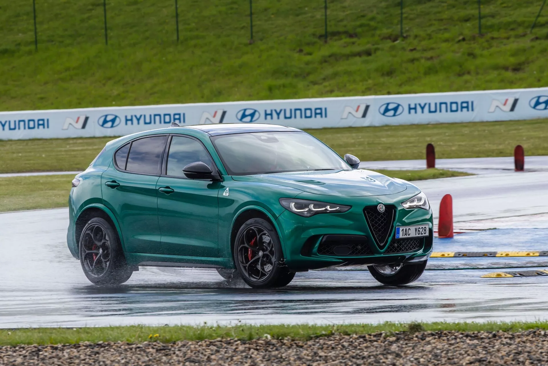 Dinamičen pogled spredaj tri četrtine na zmogljiv zelen SUV Alfa Romeo Giulia Quadrifoglio 2023 na mokri testni stezi, ki prikazuje agresiven dizajn, velika kolesa in napredne varnostne funkcije