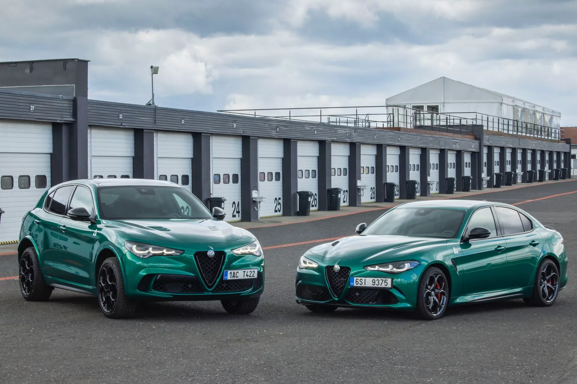 Alfa Romeo Giulia Quadrifoglio 2023 in SUV ena ob drugem v živahni zeleni, prikazujeta sprednje profile in športno oblikovanje v industrijskem ozadju z vzdušjem dirkališča, kar nakazuje na zmogljivostne funkcije, kot so LED žarometi, aerodinamične izboljšave in zavorne zmogljivosti visoke učinkovitosti.