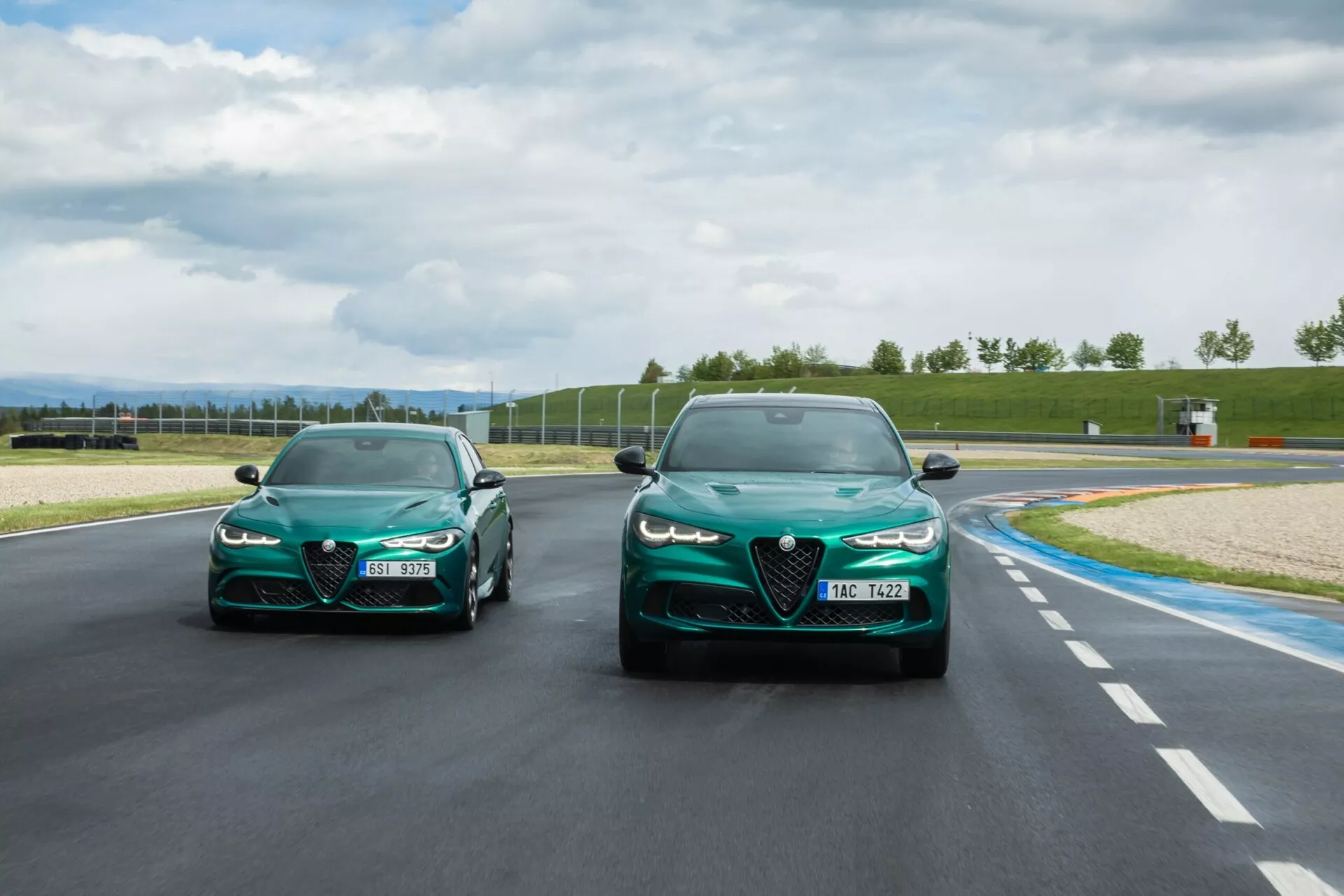 Športni avtomobili Alfa Romeo Giulia Quadrifoglio 2023 dirkajo na progi v naravni dnevni svetlobi z delno oblačnim nebom v ozadju, prikazujejo elegantne linije, aerodinamično obliko in zmogljivostne značilnosti, kot so ostri žarometi in veliki zračni vhodi