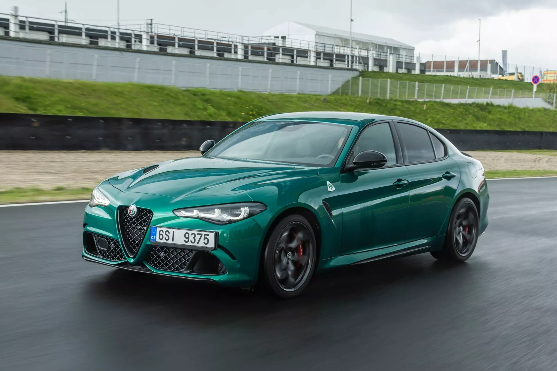 Alfa Romeo Giulia Quadrifoglio 2023 v gibanju na dirkališču, dinamičen pogled s sprednje strani v tri četrtine prikazuje agresiven slog, aerodinamično obliko, značilno masko 'Trilobe', elegantne LED žaromete, velike zračne dovode in športna grafitna kolesa z rdečimi zavornimi čeljustmi