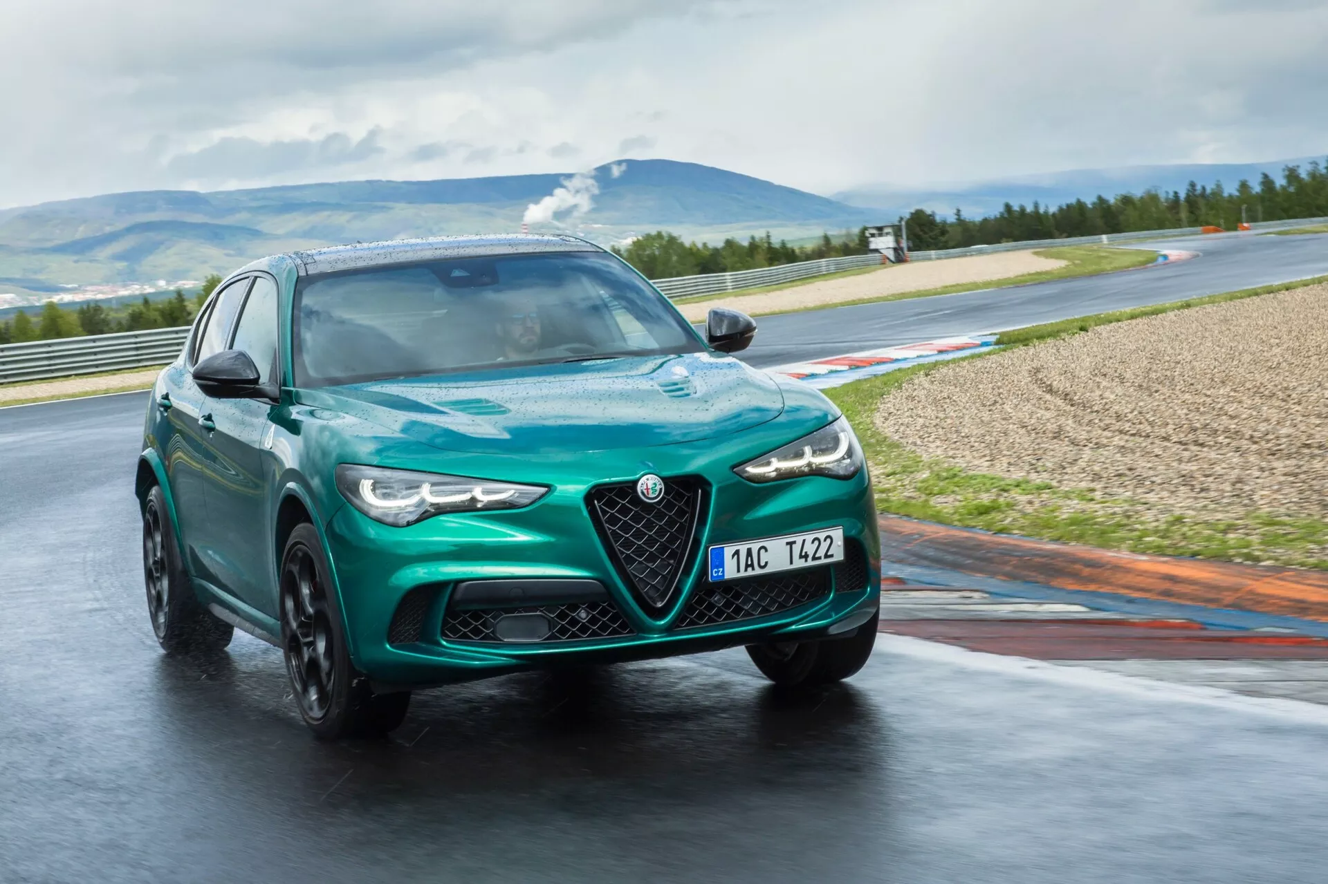Alfa Romeo Giulia Quadrifoglio 2023 v metalni zeleni, ujeta iz dinamične perspektive tri četrt spredaj na mokri dirkaški stezi, prikazuje LED žaromete, v gibanju litih koles in značilno rešetko Scudetto, na ozadju gorske pokrajine.