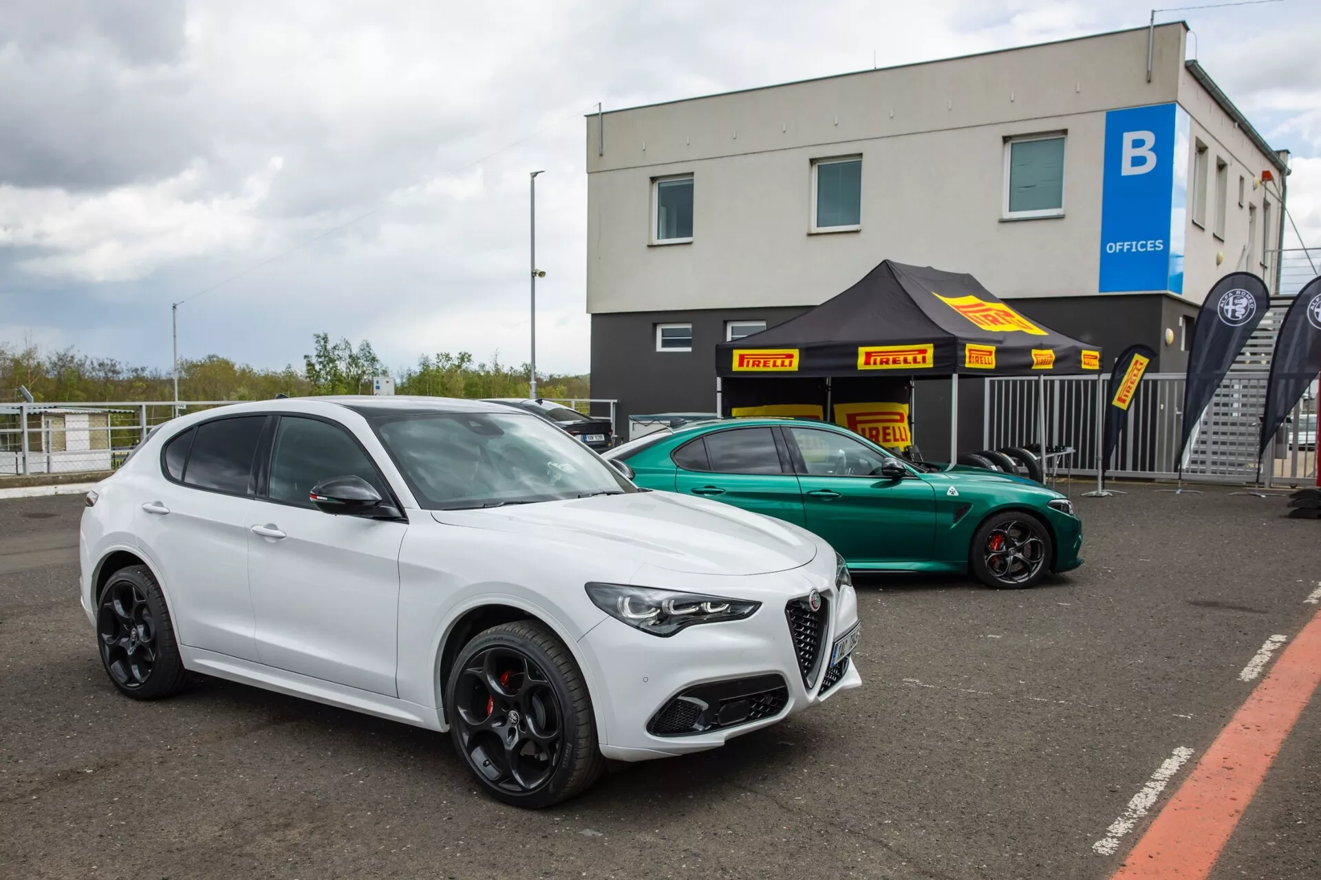Alfa Romeo Giulia Quadrifoglio 2023 in luksuzni SUV parkirana na asfaltirani površini v potencialni dirkališči ali industrijskem kompleksu pisarniškega območja, ki prikazuje značilno značkino značko, na zmogljivost usmerjena črna kolesa in agresiven styling v oblačni dnevni svetlobi