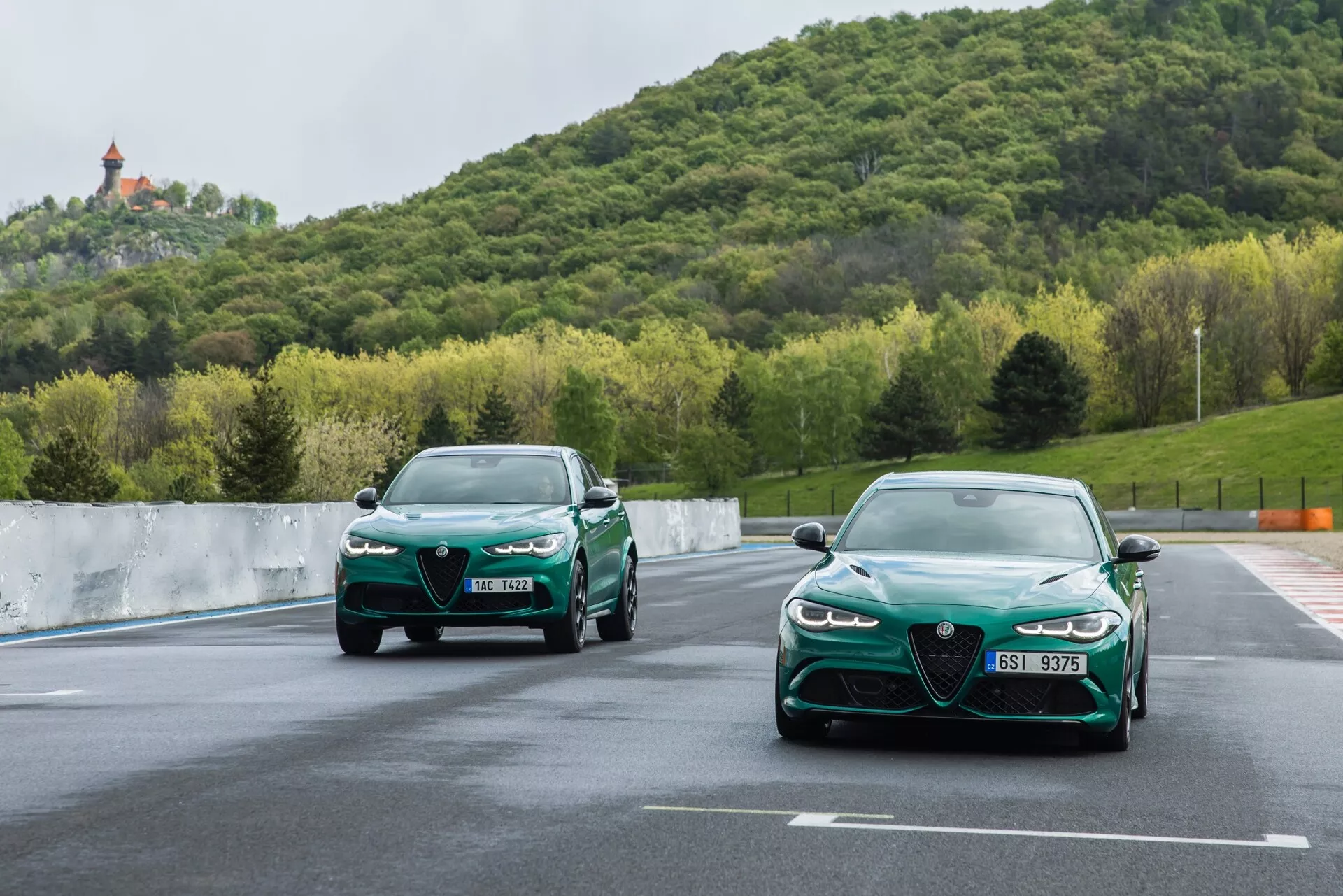 Dinamičen pogled na dva avtomobila Alfa-Romeo Giulia Quadrifoglio 2023, ki se vozita po stezi, s podrobnim fokusom na sprednjo rešetko, LED žaromete in aerodinamično zasnovo na ozadju zelenja in hriba pod oblačnim nebom