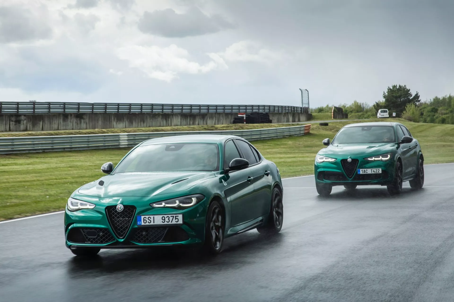 Alfa Romeo Giulia Quadrifoglio 2023 v metalik zeleni, prikazujejoč svojo veliko značilno rešetko, LED žaromete in športna lita kolesa na mokri progi, poudarjajoč napredne zmogljivostne in varnostne funkcije
