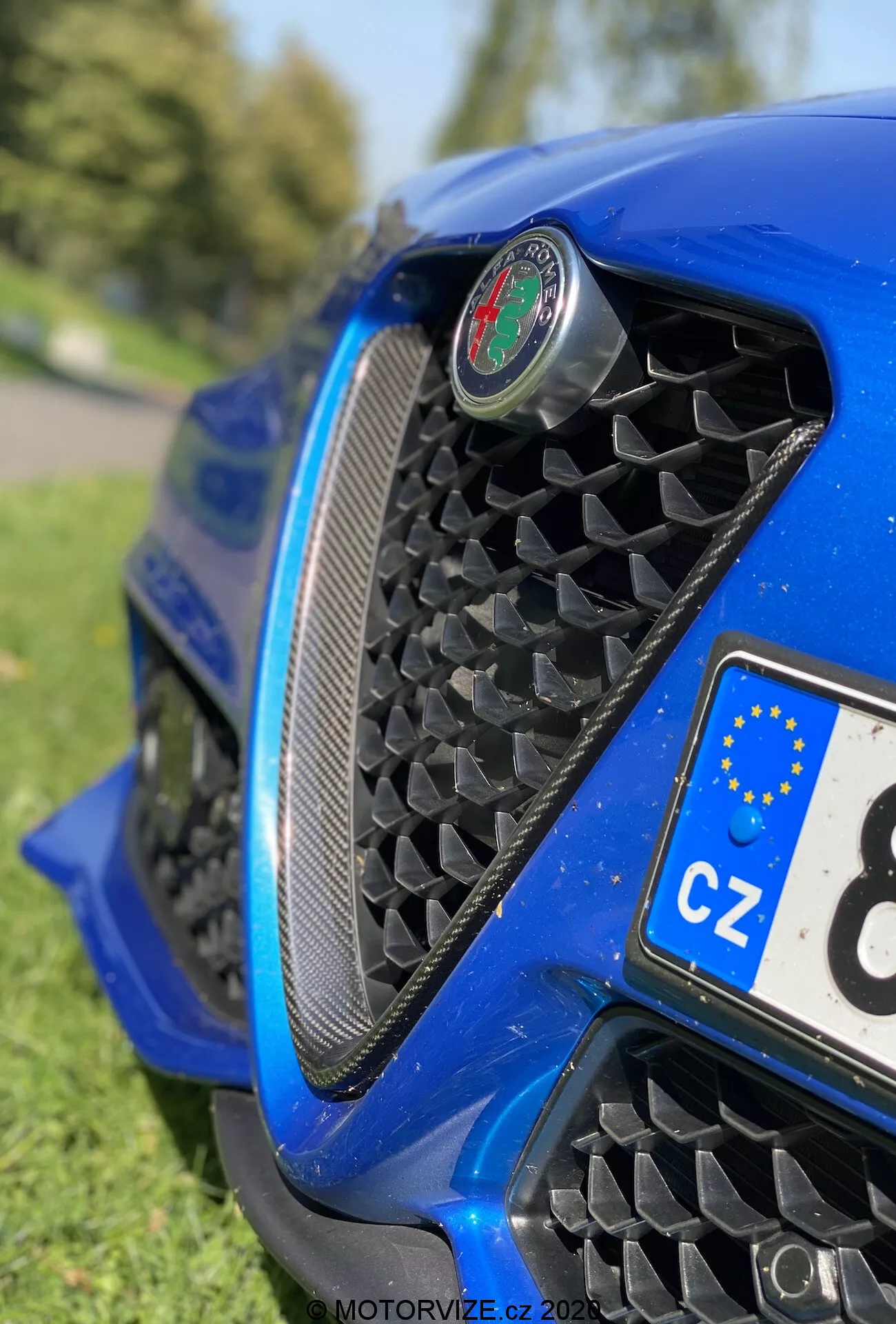 Pogled od blizu na sprednji del Alfa Romeo Giulia 2016 iz nizkega kota, ki poudarja mrežo v obliki satja, osrednji emblem, aerodinamičen sprednji odbijač in sijočo kovinsko modro karoserijo z odsevi. Delni pogled na češko registrsko tablico in zamegljeno okolje, podobno parku.