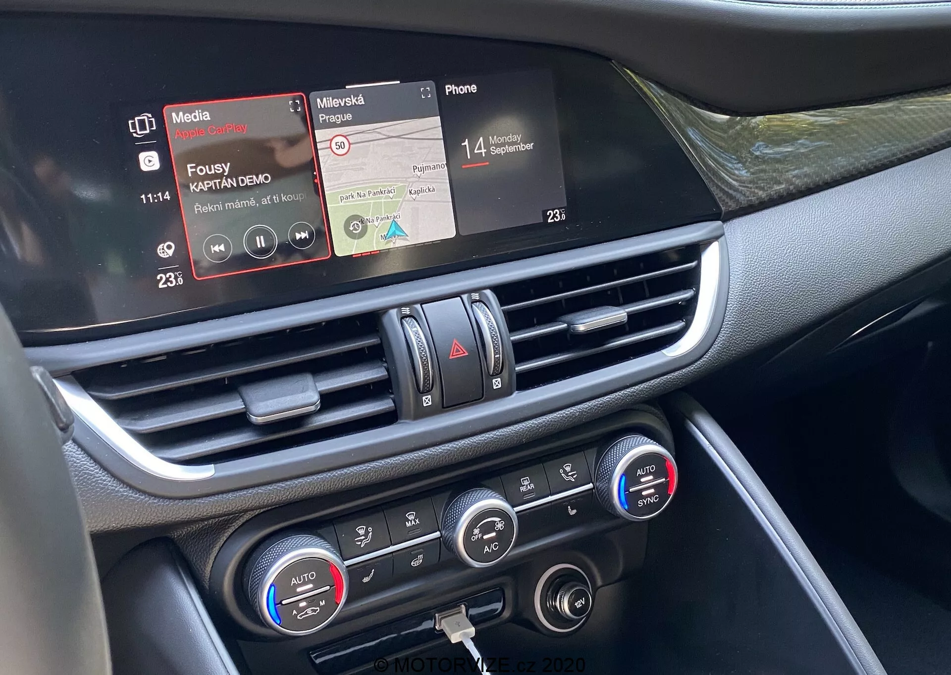 Bližnji posnetek osrednjega dela armaturne plošče Alfa Romeo Giulia 2016 iz perspektive sprednjega sovoznika. Srednji bližnji posnetek poudarja infotainment zaslon na dotik s funkcijami predvajalnika medijev in povezljivosti s telefonom, osrednji zračni izpust s kromiranimi poudarki in nadzorne elemente klimatske naprave z nastavitvami temperature za dve območji ter digitalne prikazovalnike. Materiali armaturne plošče se zdijo izdelani iz plastike ter kovinskih ali kromiranih elementov, kar prikazuje napredno notranjo tehnologijo vozila, osredotočeno na povezljivost, predvajanje medijev in nadzor klimatske naprave.