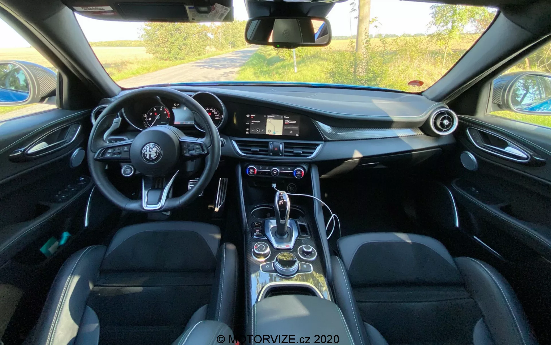 Pogled na notranjost Alfa Romeo Giulia 2016 iz perspektive voznika, ki prikazuje volan s plosko dno z več upravljalnimi gumbi, digitalni informacijski sistem na armaturni plošči, analogno uro, okrogle zračnike, elegantno prestavno ročico, izbirnik načinov vožnje, držala za kozarce, električno upravljane okenske sisteme, usnjene sedeže s stranskimi oporniki in splošne sodobne tehnološke funkcije.