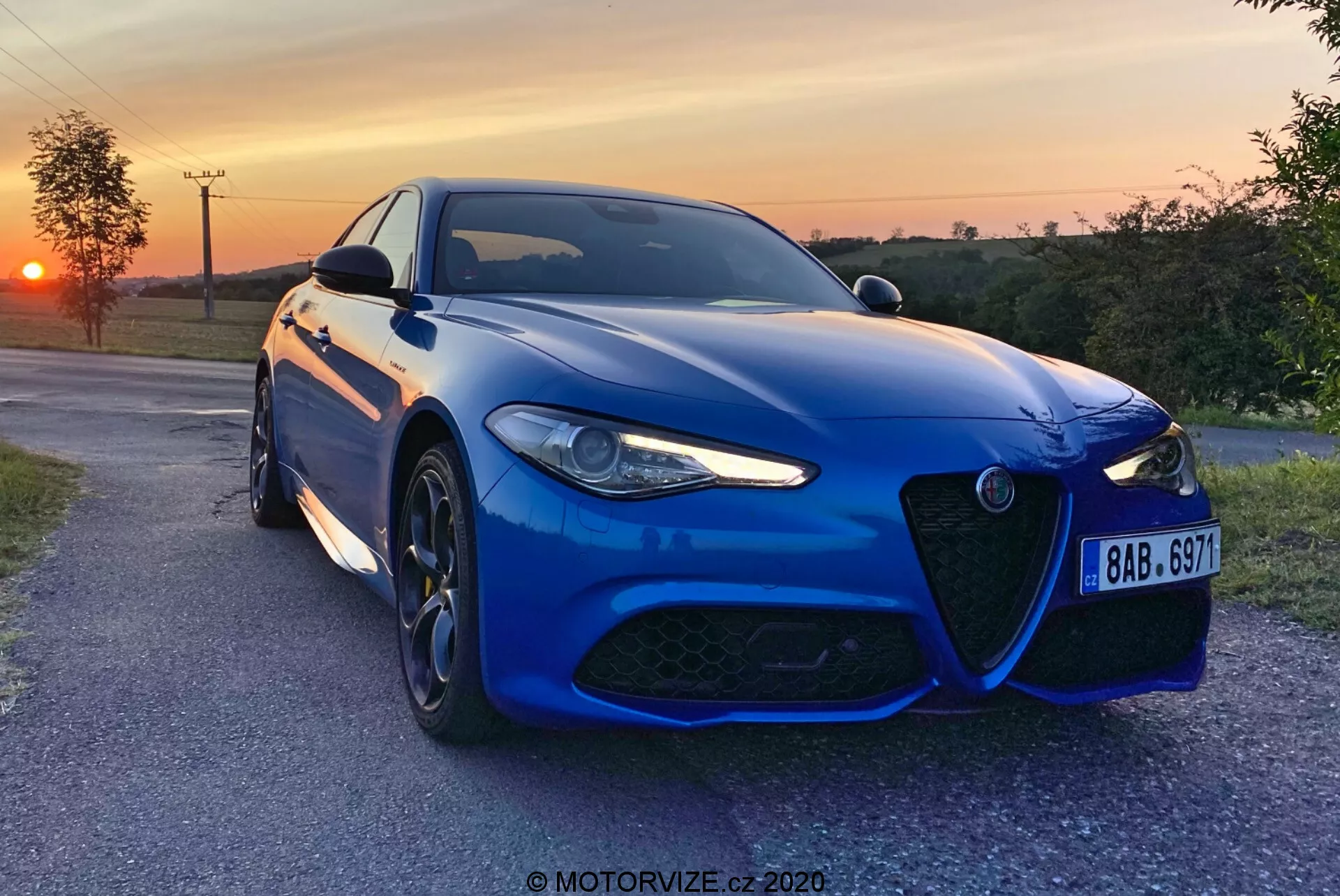 Moder športni limuzini Alfa-Romeo Giulia 2016, posnet zvečer s sončnim zahodom v ozadju, postavljen rahlo pod kotom proti fotoaparatu na tlakovanem podeželski cesti, prikazuje svojo značilno sprednjo masko, agresiven dizajn LED žarometov, velika večkraka platišča iz lahke litine in visokozmogljiv zavorni sistem. Kovinska barva, aerodinamičen sprednji odbijač, izklesane linije pokrova motorja in napredni senzorji za asistenčne sisteme voznika poudarjajo športno estetiko in sodobno tehnologijo avtomobila. Prisotna je vodna oznaka 'MOTORVIZE.cz 2020'.