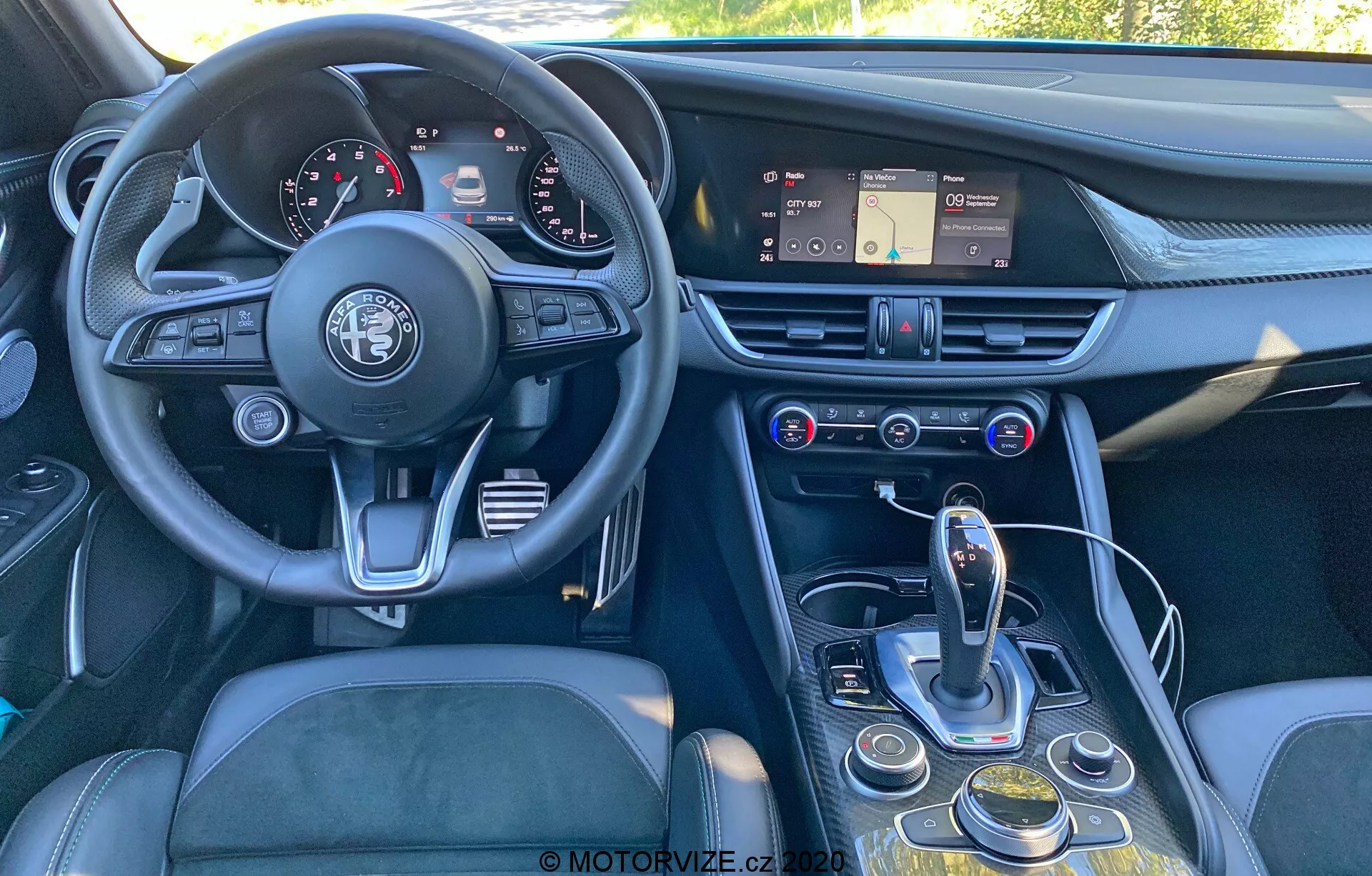 Pogled na notranjost Alfa Romeo Giulia iz leta 2016 iz rahlo premaknjenega kota od zadaj naprej, ki prikazuje armaturno ploščo, volan, sredinsko konzolo in del voznikovega sedeža. Slika poudarja trikraki volan z emblejem Alfa Romeo, instrumentno ploščo z analognimi merilniki in digitalnimi zasloni, armaturno ploščo z zaslonom na dotik za infotainment, zračniki in kontrolami za klimo in multimedijo. Sredinska konzola vključuje avtomatski izbirnik prestav, vrtljivi gumb, gumbe za navigacijo infotainmenta in nastavitve vozila ter možen izbirnik načina. Oblikovanje poudarja ergonomijo in dostopnost, z materiali, kot so usnje, brušena kovina in visoko kakovostna plastika, ki dajejo občutek luksuza.