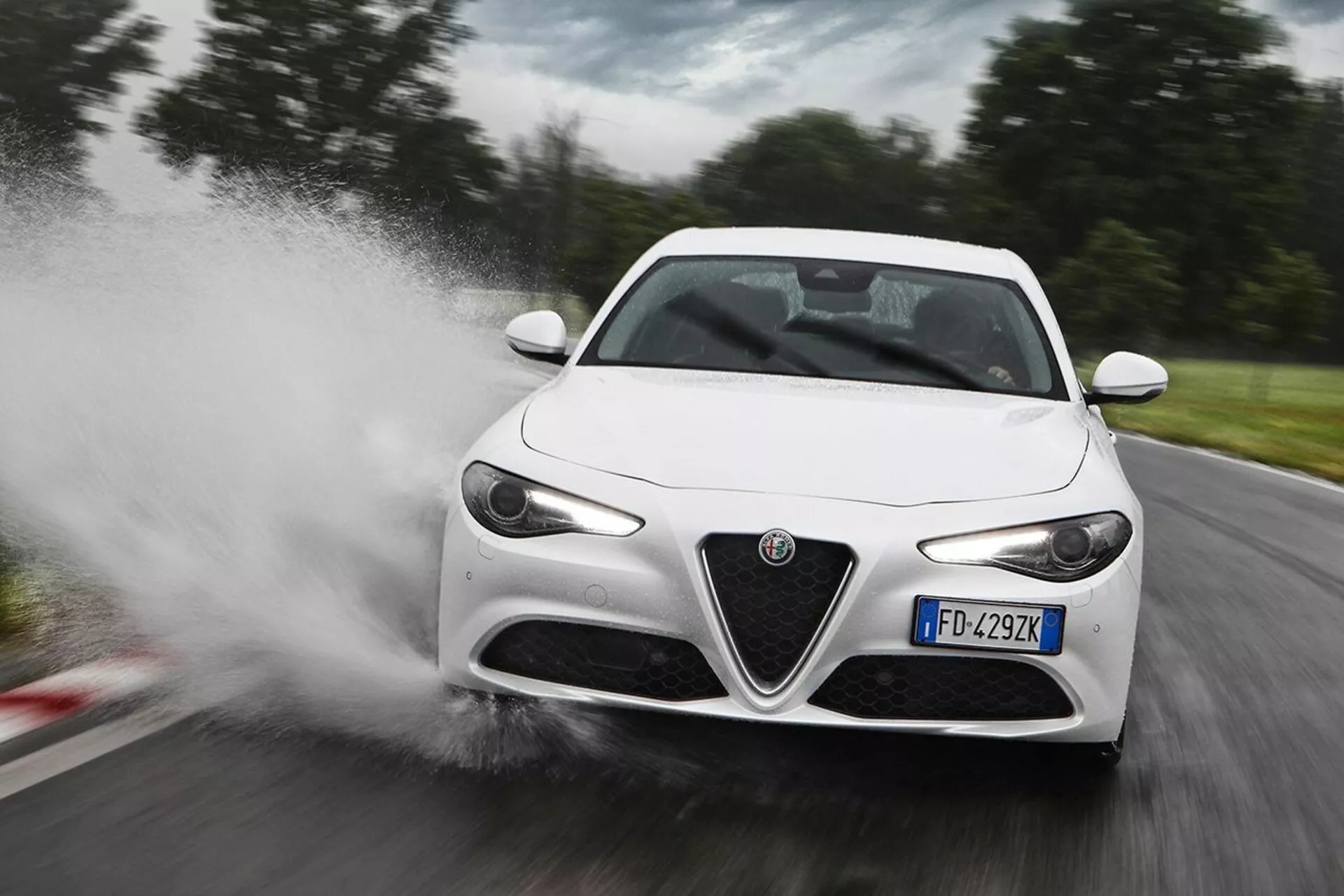 Beli športni avtomobil Alfa Romeo Giulia iz leta 2016, ki z visoko hitrostjo prečka vodo in povzroča škropljenje, v pogledu od spredaj tri četrtine, ki poudarja njegove LED žaromete, značilno sprednjo masko, elegantna stranska ogledala in aerodinamične linije