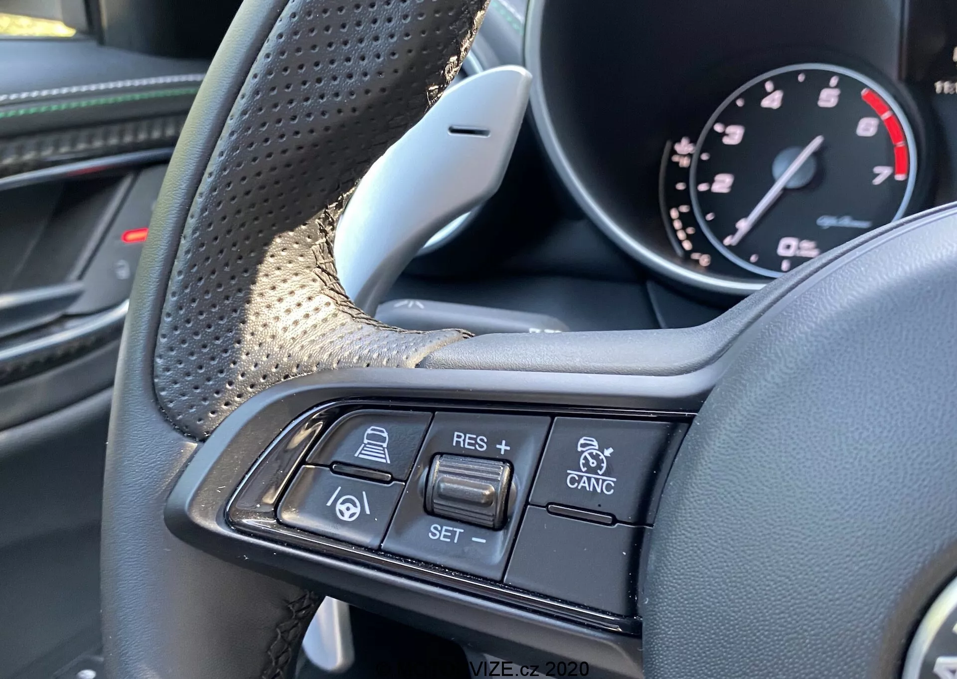 Pogled od blizu notranjosti Alfa-Romeo Giulia 2016, ki prikazuje volan in njegove večfunkcijske kontrole iz perspektive voznika. Na levi strani so vidna teksturirana prijemališča in luksuzni materiali, kot sta usnje in morda karbonska vlakna. Desno krak volana vsebuje gumbe za tempomat in prilagodljivo tehnologijo tempomata, vključno z + RES, - SET, CANCEL, ikono avto-sledi-avto in opozorila o koliziji vozil. Sliko se osredotoča izključno na te kontrole, poudarja premium dizajn in napredne funkcije za pomoč vozniku.