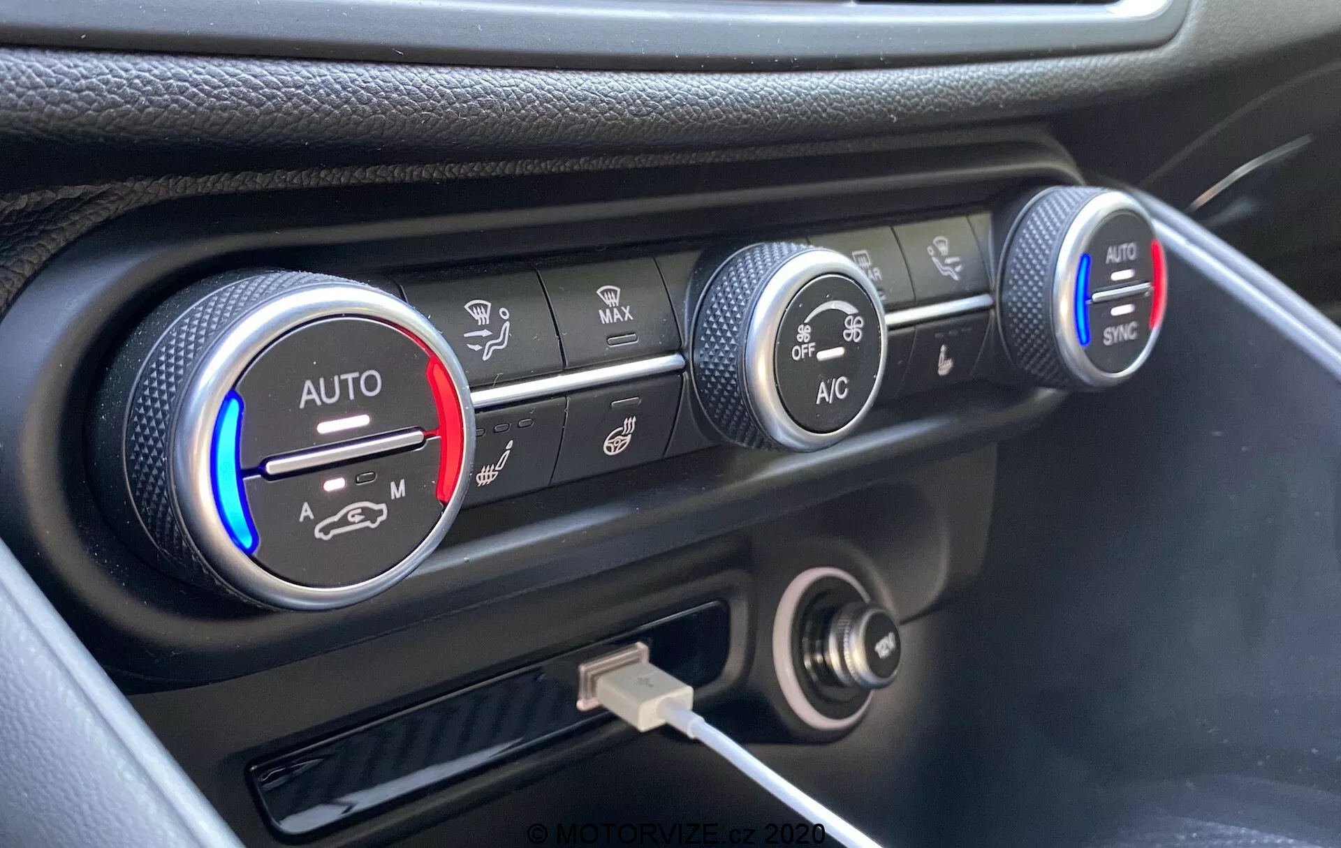 Podroben pogled na klimatski sistem na armaturni plošči Alfa Romeo Giulia 2016, s tremi krožnimi gumbi. Levi gumb označuje nastavitve temperature z modrimi in rdečimi trakovi ter nastavitvijo 'AUTO', delno osvetljen z rdečo. Srednji gumb prikazuje nastavitve hitrosti ventilatorja, kar je poudarjeno pri nižji hitrosti. Desni gumb prikazuje različne načine porazdelitve zraka s tipkama 'AC' in 'SYNC'. Pod gumbi je 12-voltna vtičnica in USB priključek. Armaturna plošča je črne barve z srebrnim poudarkom okoli klimatskih kontrol, posneta iz dvignjenega kota.