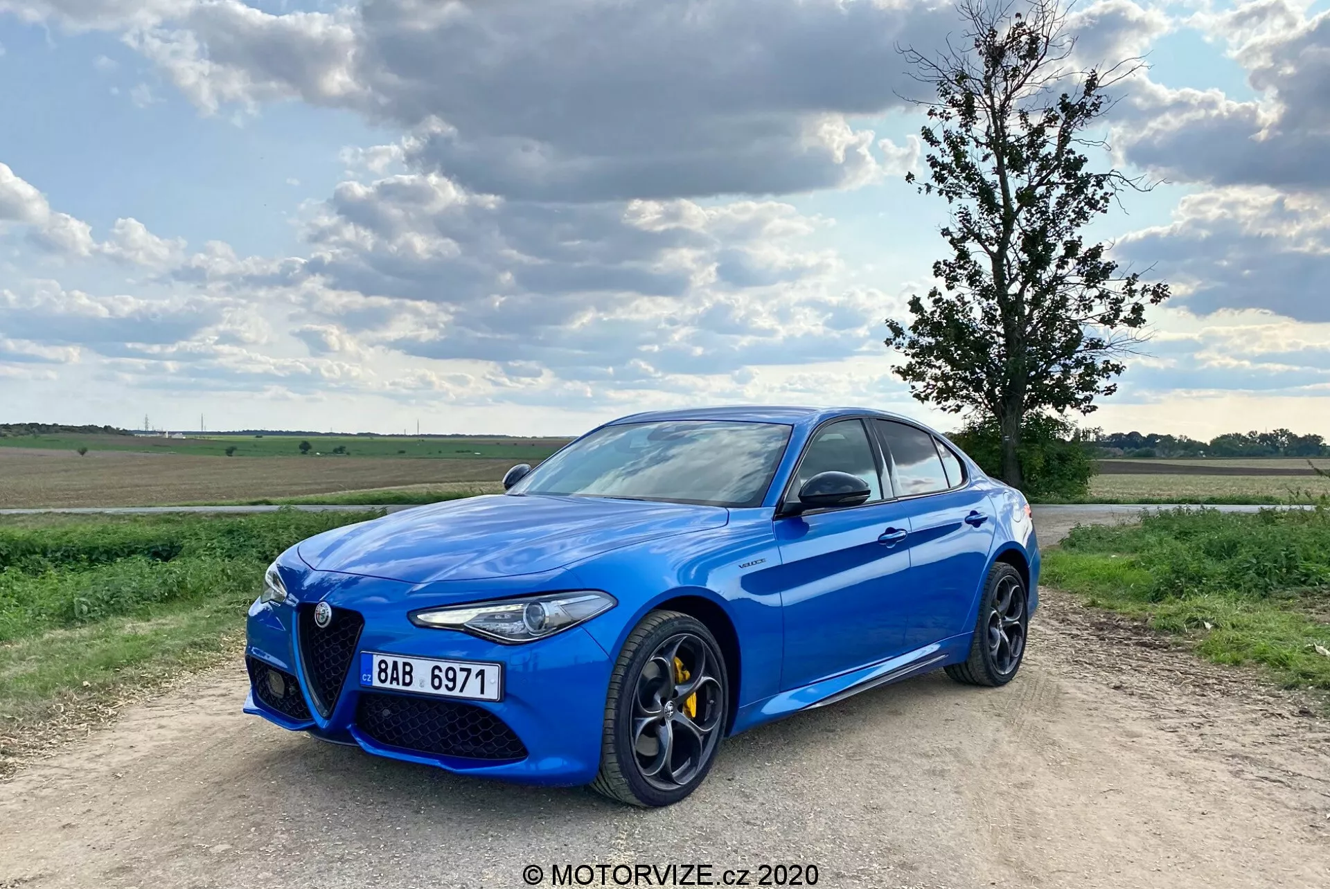 Slika svetlo modrega športnega sedana Alfa Romeo Giulia 2016 parkiranega na makadamski poti z delno oblacnim modrim nebom v ozadju. Avto je ujet iz triosminskega sprednjega kota, kar prikazuje njegovo značilno prednjo masko 'Trilobo', rumene zavorne čeljusti, velike večkrake aluminijaste platišča, elegantne LED žaromete in kromirane okenske okvire. Vozilo je nekoliko levo od sredine s sprednjim levim kolesom, ki kaže navzven, kar mu daje dinamično držo.