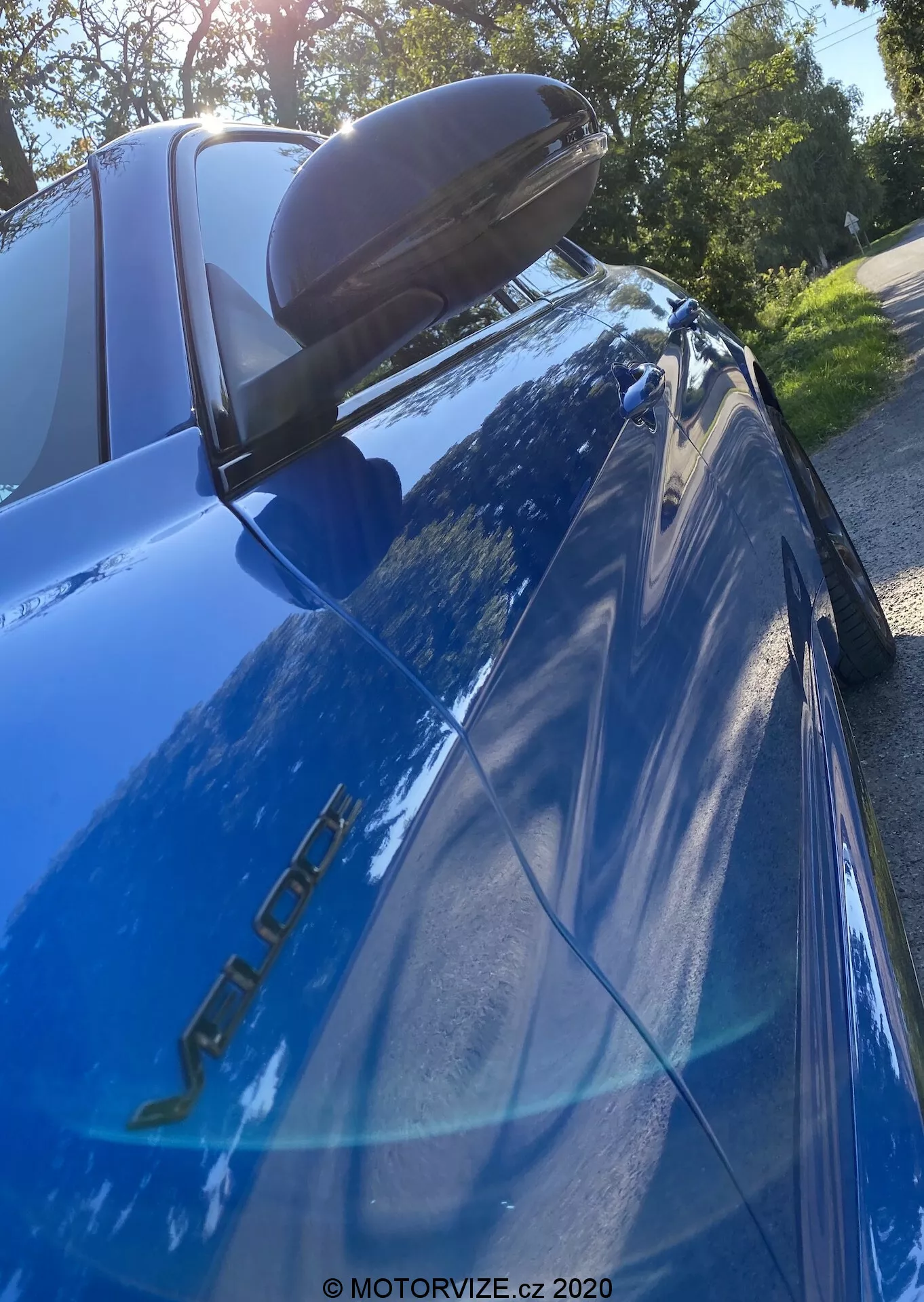 Posnetek od blizu modrega Alfa Romeo Giulia (model 2016) fotografiran iz nizkega in navzgor usmerjenega kota s poudarkom na zadnjem delu. Vidni so znaki modela na strani voznika, odsevna modra barva, levo zadnje LED luči, črn zadnji spojler, padajoče zadnje okno s tretjo zavorno lučjo, odsev dreves in svetlega neba ter stransko ogledalo z integriranim smernikom.