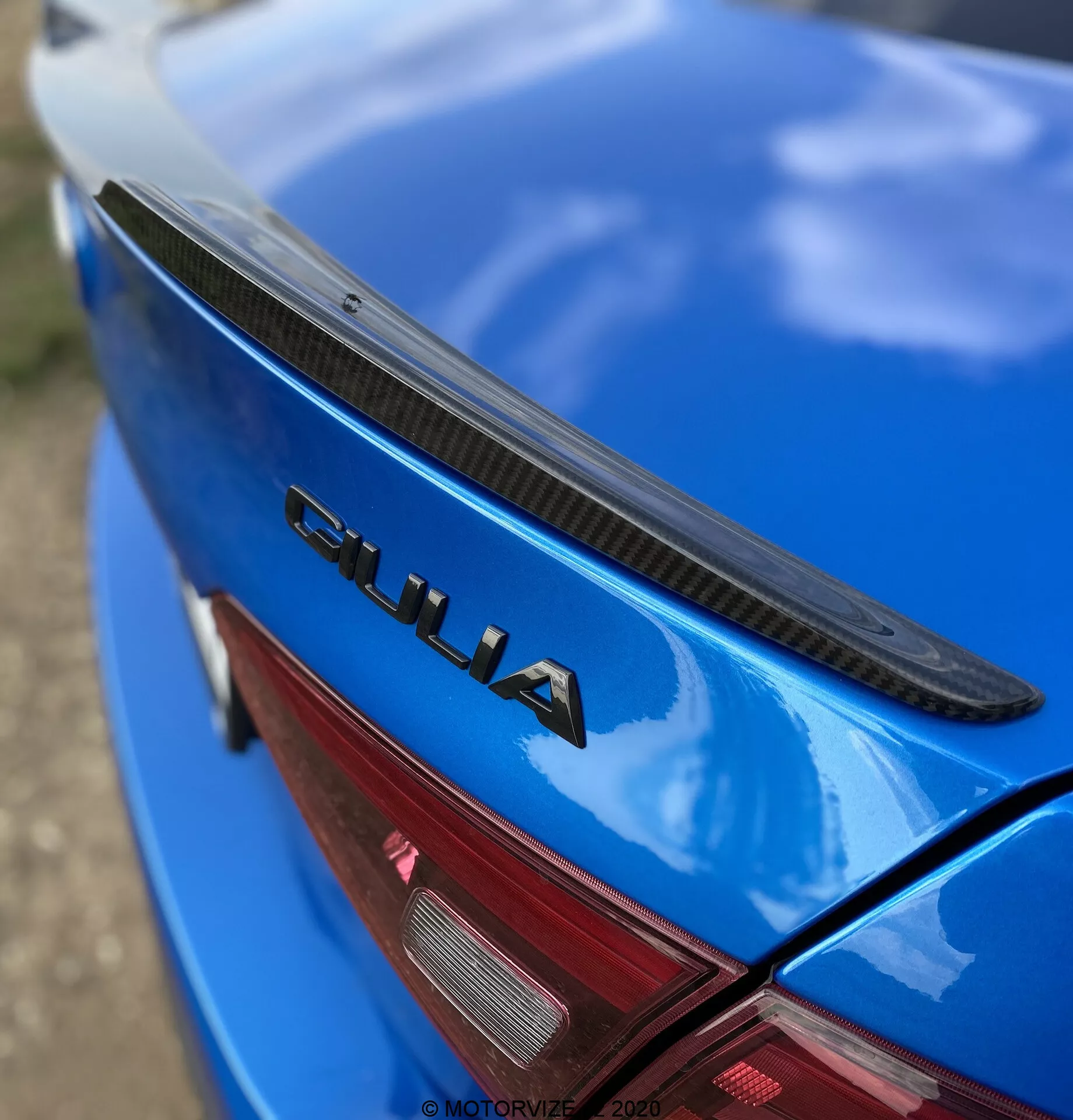 Podroben, poševen pogled na zadnji del modrega Alfa Romeo Giulia 2016. Fotografija prikazuje pokrov prtljažnika s črnim spojlerjem, sijočo modro barvo in oznako 'GIULIA', ki je dobro vidna v kromu. Vidni so tudi del rdeče LED zadnje luči. Oznaka 'GIULIA' je ostro izostrena, medtem ko se spojler, površina avtomobila in zadnja luč postopoma zameglijo, kar poudarja elemente znamke in oblikovanja avtomobila.