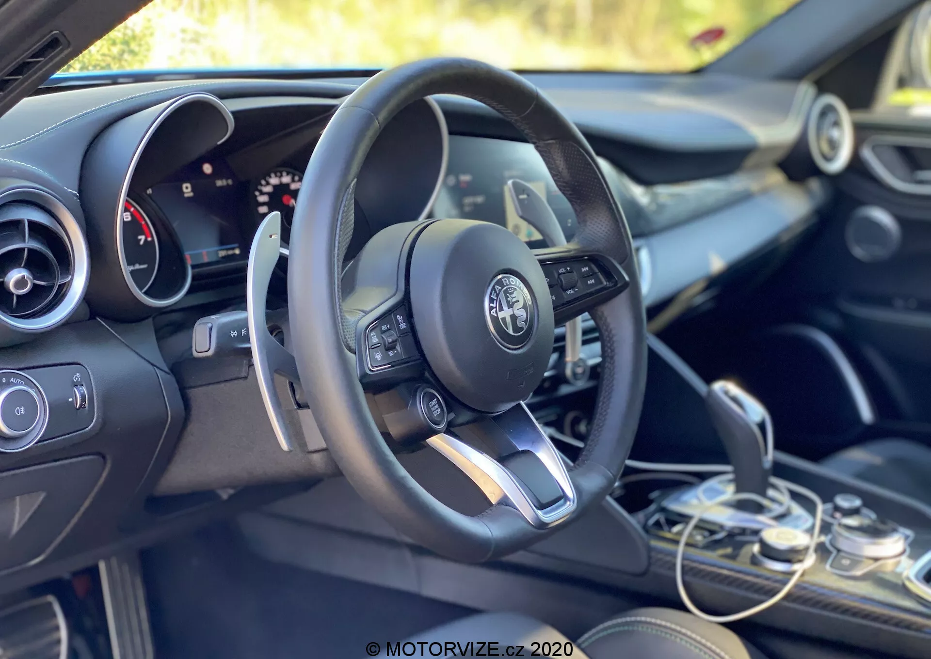 Notranji pogled na alfa romeo giulia 2016, osredotočen na voznikov prostor in armaturno ploščo z leve strani, rahlo proti zadnji strani. Konfiguracija za levo roko prikazuje moderen, večfunkcijski volan z vgrajenimi kontrolami, vključno z gumbi za uravnavanje glasnosti, dostop do menijev in verjetno tempomat. Logotip Alfa Romeo je jasno viden na sredini volana. Za volanom je delno viden kombinirani analogni in digitalni prikazovalnik, ki nakazuje na konfigurabilen informacijski sistem za voznika. Na desni strani volana je rahlo viden zgornji del zaslona infotainment sistema, spodaj pa so klimatski sistemi in funkcionalna stikala. Menjalna ročica se nahaja v spodnjem desnem kotu in prikazuje eleganten dizajn avtomatskega menjalnika. Notranjost združuje temne odtenke z kovinskimi poudarki, kar odraža prestižen dizajn vozila. Kabina je obsijana z naravno svetlobo, zelena kulisa pa nakazuje zunanje ali vegetacijsko okolje.