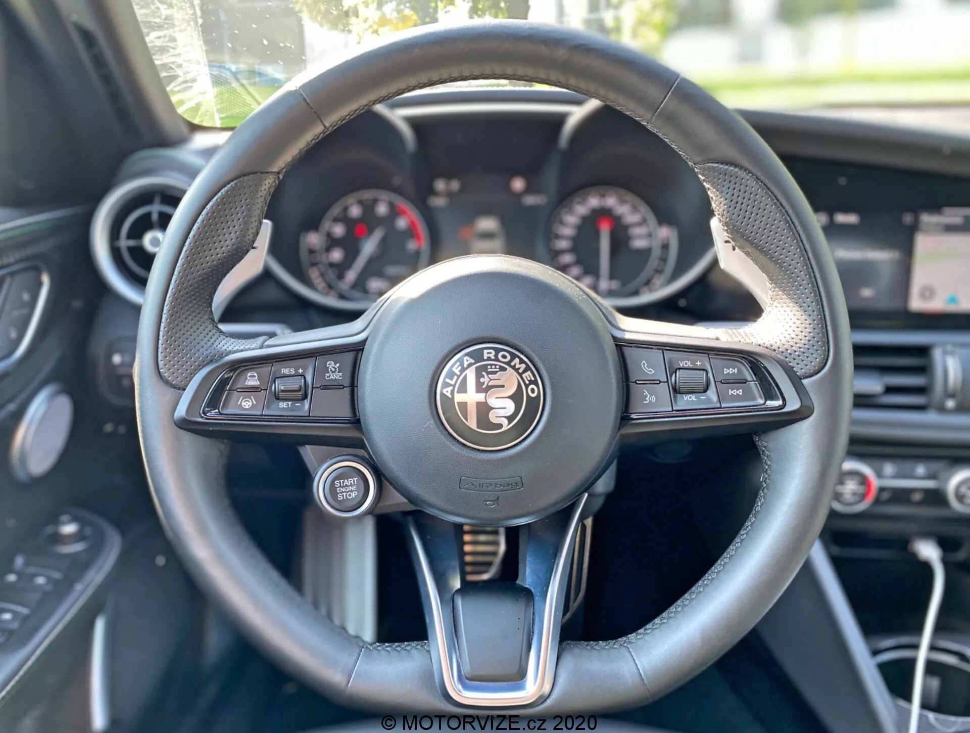 Voznikov pogled na notranjost Alfa-Romeo Giulia 2016, osredotočen na volan in armaturno ploščo. Centralno nameščen multifunkcijski volan ima gumbe za medije, glasovno upravljanje, telefonsko povezljivost, informacije o vozilu in tempomat. Instrumentna plošča prikazuje analogne merilnike, verjetno merilnik hitrosti in obratomer, z digitalnim zaslonom vmes. Volan združuje usnje, šive in kovinske poudarke, kar daje občutek razkošja. V ozadju so vidni gumbi za nadzor klime in nad njimi osrednji zaslon.
