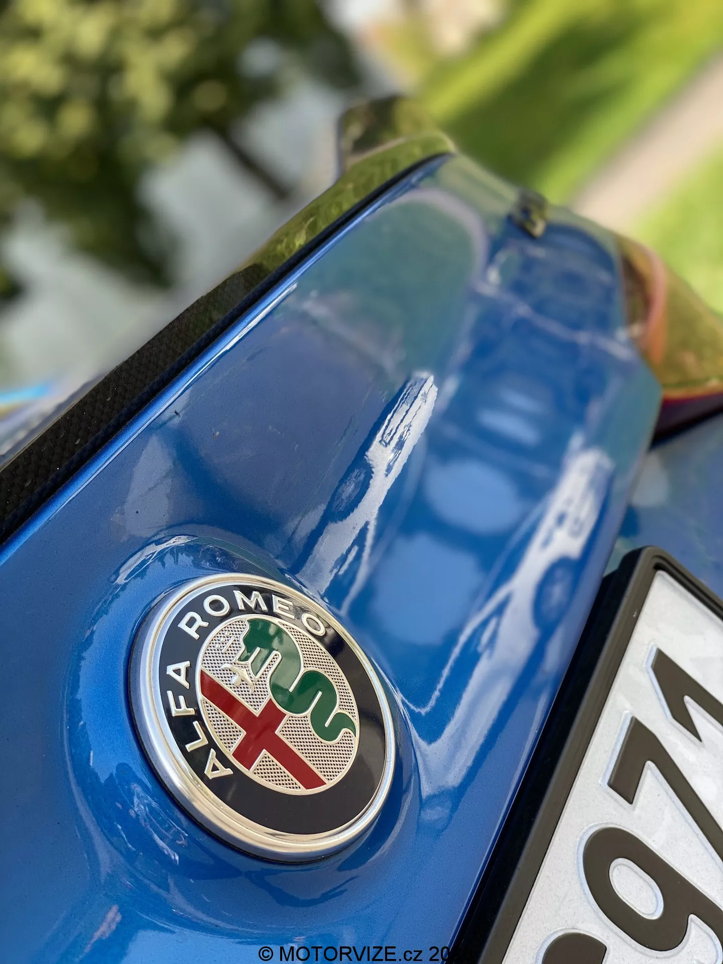 Približan pogled na zadnji del Alfa Romeo Giulia 2016 s poudarkom na emblemu blagovne znamke iz nizkega kota. Znak, s rdečim križem na belem ozadju in zeleno kačo na modrem ozadju, je jasno izostren na kovinsko modrem pokrovu prtljažnika s podrobnostmi in odsevi okoliške površine.