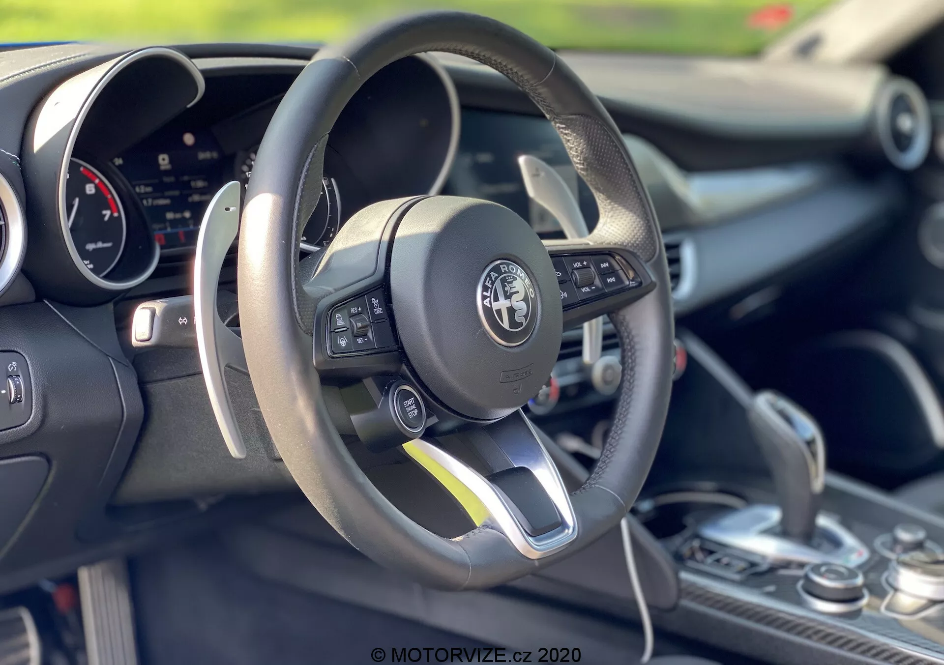 Posnetek notranjosti Alfa-Romeo Giulia iz leta 2016, ki se osredotoča na območje voznika in volan. Ploščat spodnji del, usnjen volan vsebuje večfunkcijske kontrole na levi strani. Za volanom je viden digitalni instrumentni zaslon. Sredinska konzola prikazuje prestavno ročico z usnjenim ovojem, kar nakazuje na možnost ročnega prestavljanja. Slika poudarja športno zasnovo avtomobila, napredno tehnologijo in podrobno obrtniško izdelavo z majhno globinsko ostrino, ki poudarja elemente v ospredju.