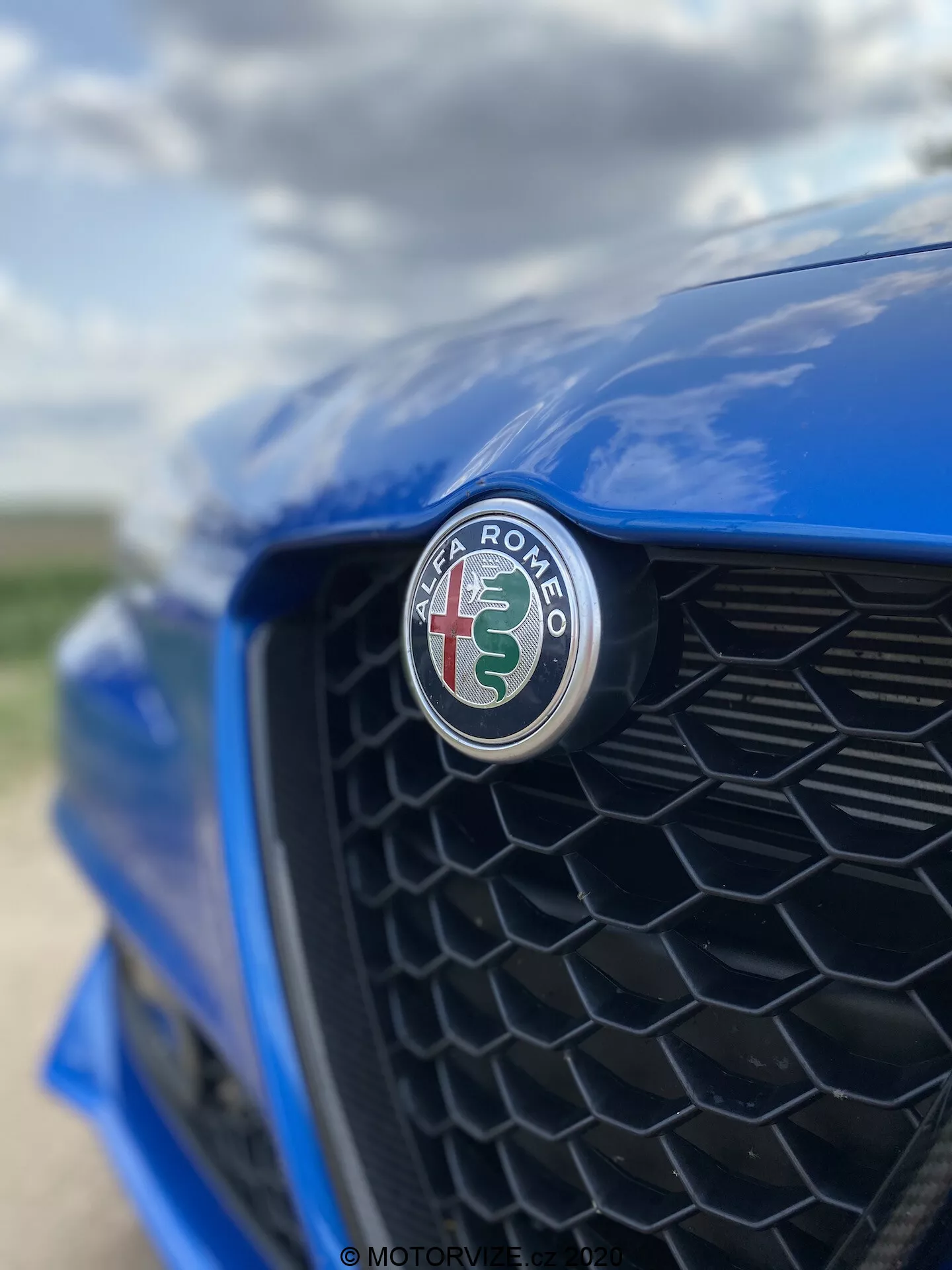 Povečava sprednjega dela modrega avtomobila Alfa Romeo Giulia 2016, s poudarkom na emblem izdelovalca z rdečim križem in zeleno kačo na belem ozadju. Posnetek iz zunajcentričnega kota proti voznikovi strani, črna plastična rešetka je rahlo zamegljena, kar poudarja ostri fokus emblema in sijočo površino.