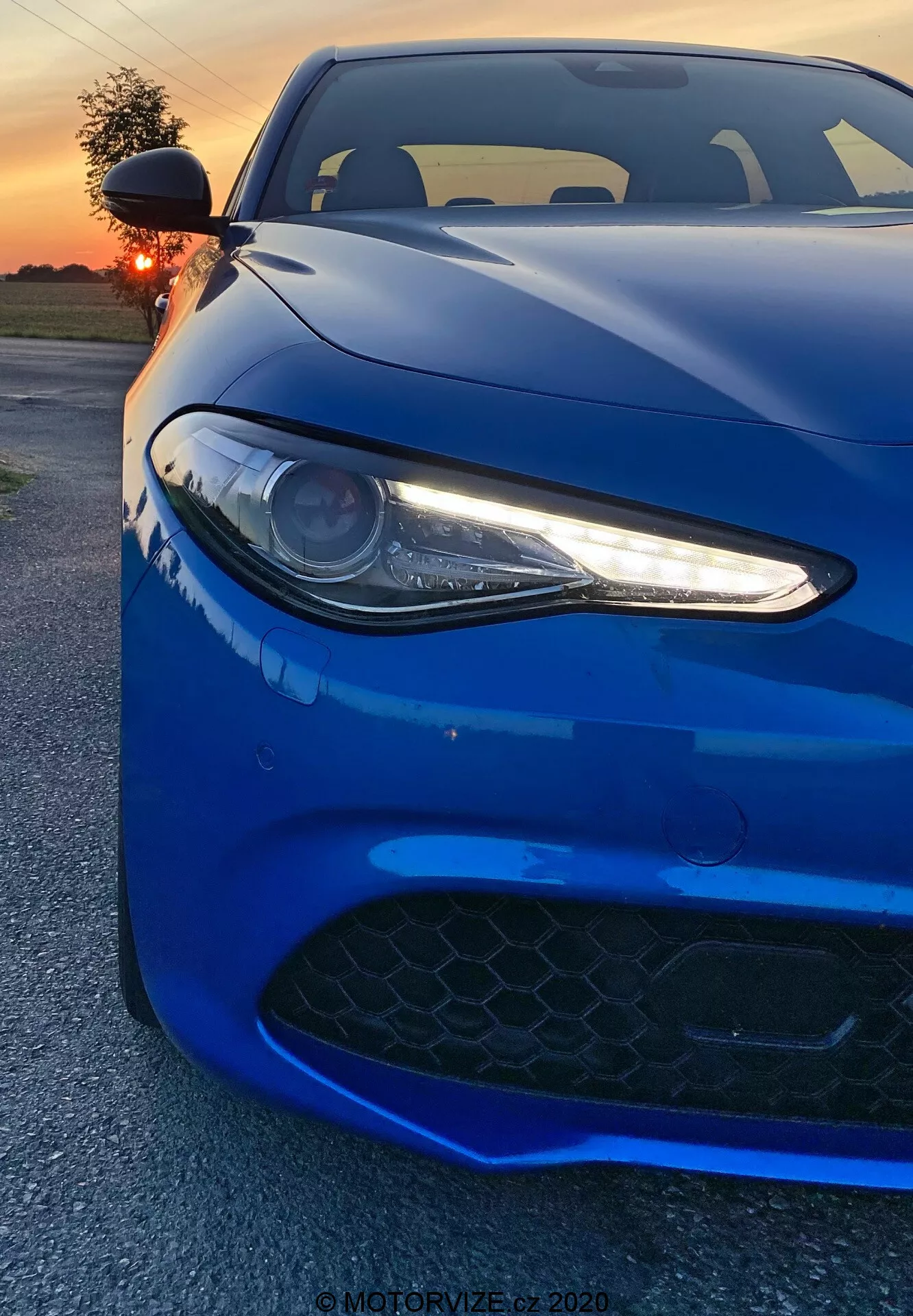 Slika prikazuje športni avtomobil Alfa Romeo Giulia 2016, posneti iz nižjega sprednjega kota. Vibrantna temno modra vozilo zapolni okvir, poudarjajoč levi žaromet z LED dnevnimi lučmi, del pokrova motorja in rešetko za dovod zraka v sprednjem odbijaču. Projektorski žaromet s tankimi belimi LED poudarki dodaja agresiven, moden videz. Črna rešetka s šesterokotnim vzorcem nakazuje visoke zmogljivosti. Posnetek zajema aerodinamične linije in obrise avtomobila, z odsevi zahajajočega sonca, ki poudarjajo sijajno površino. V ozadju je vidno toplo oranžno nebo, delno skrito za soncem. Fotografov bližnji kot poudarja oblikovne podrobnosti, ki vzbujajo občutek moči in elegance.